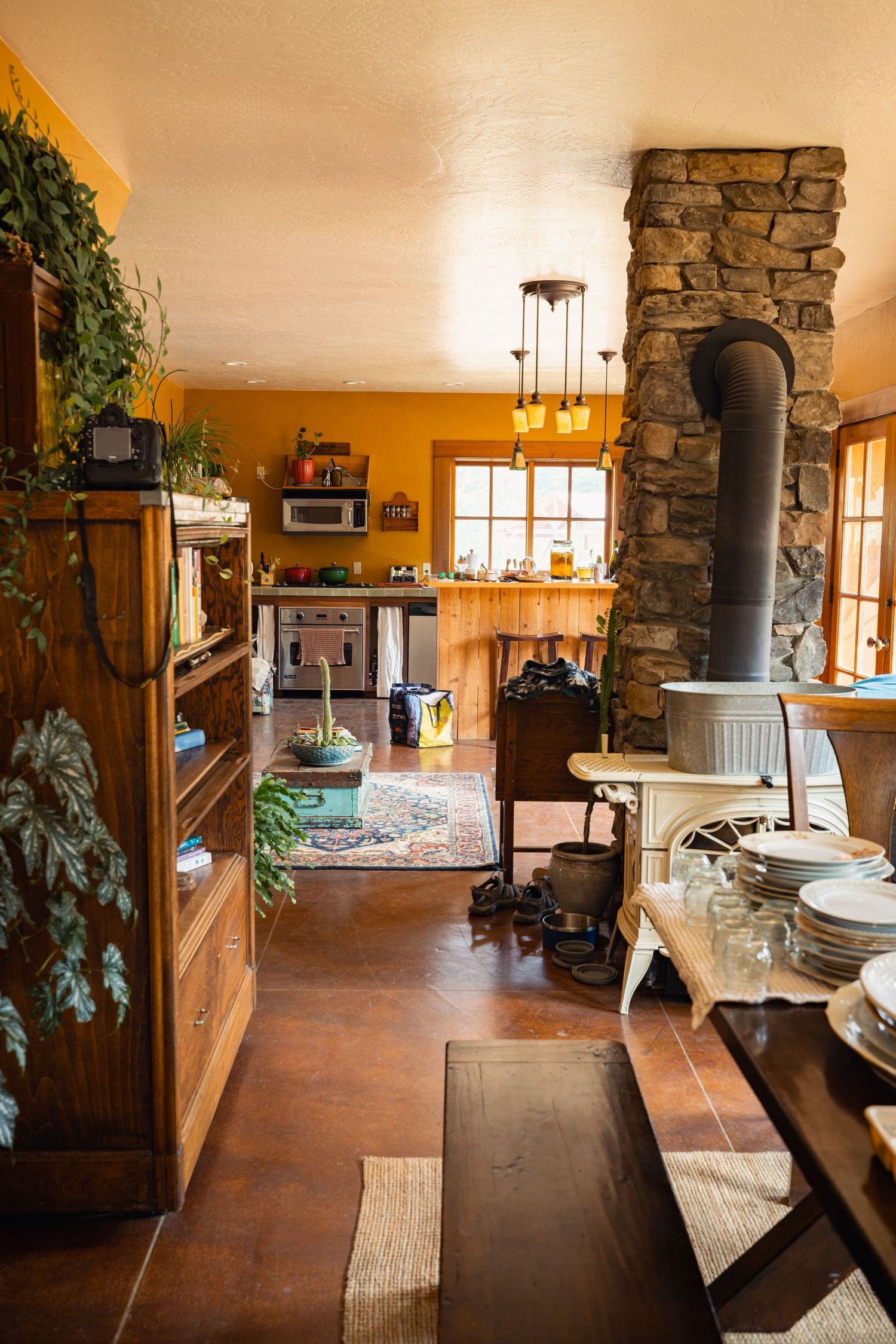 Cozy rustic kitchen interior with stone fireplace, wooden floors, yellow walls and hanging pendant lights.