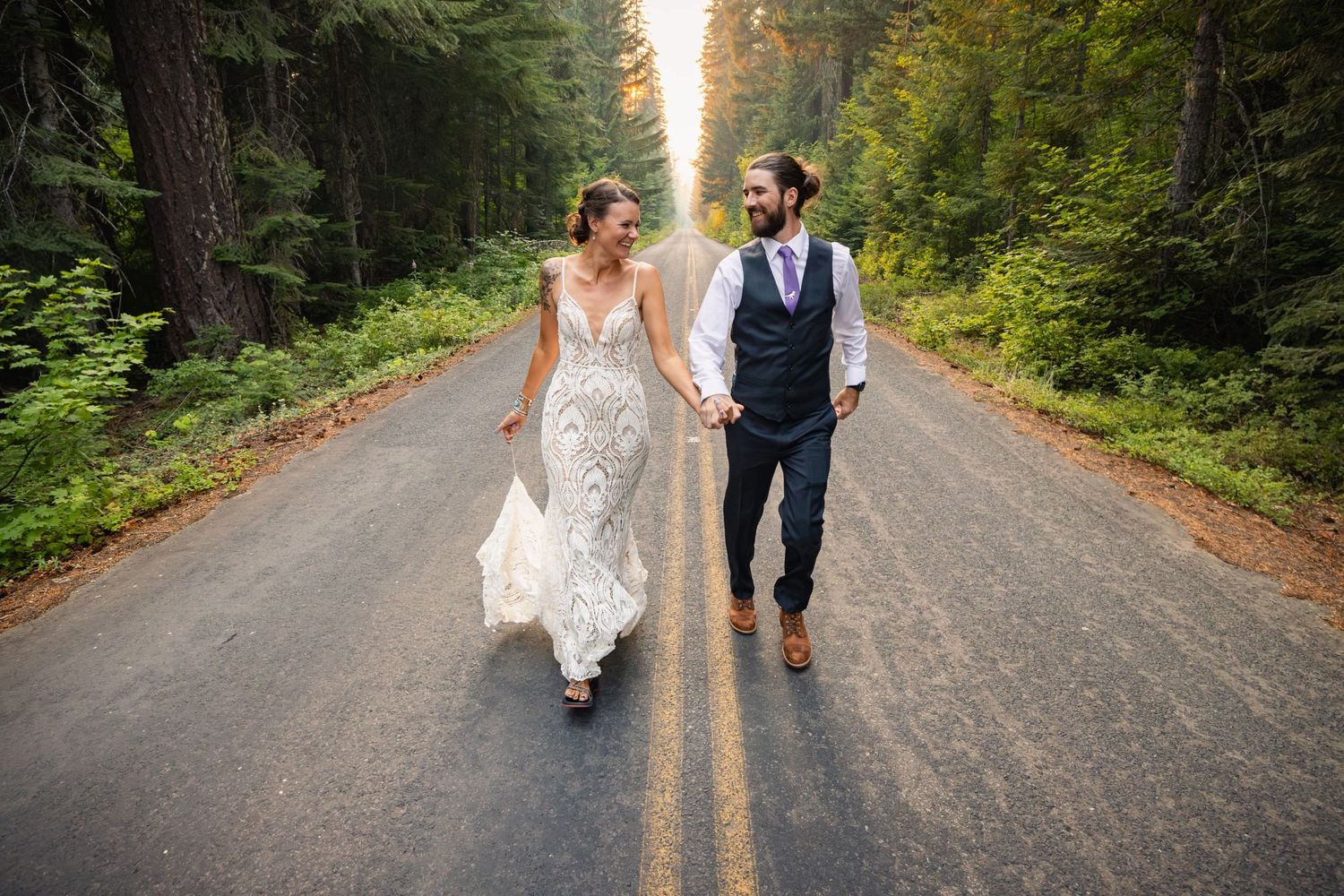A couple in wedding attire walks down a tree-lined forest road on their special day.