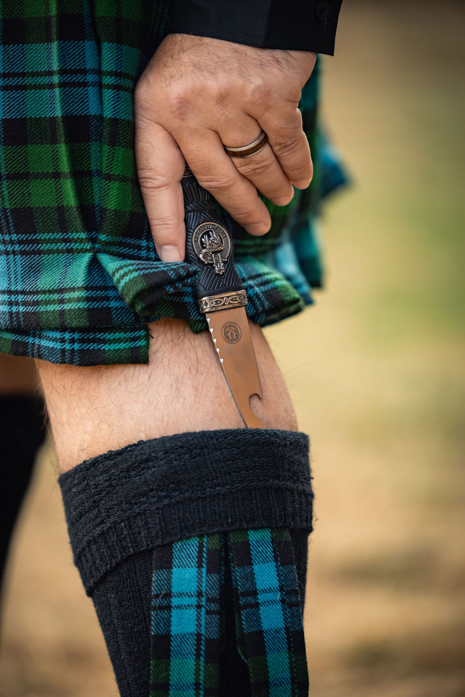 Close-up of a hand holding an ornate sgian-dubh knife against traditional green and blue tartan kilt and socks.