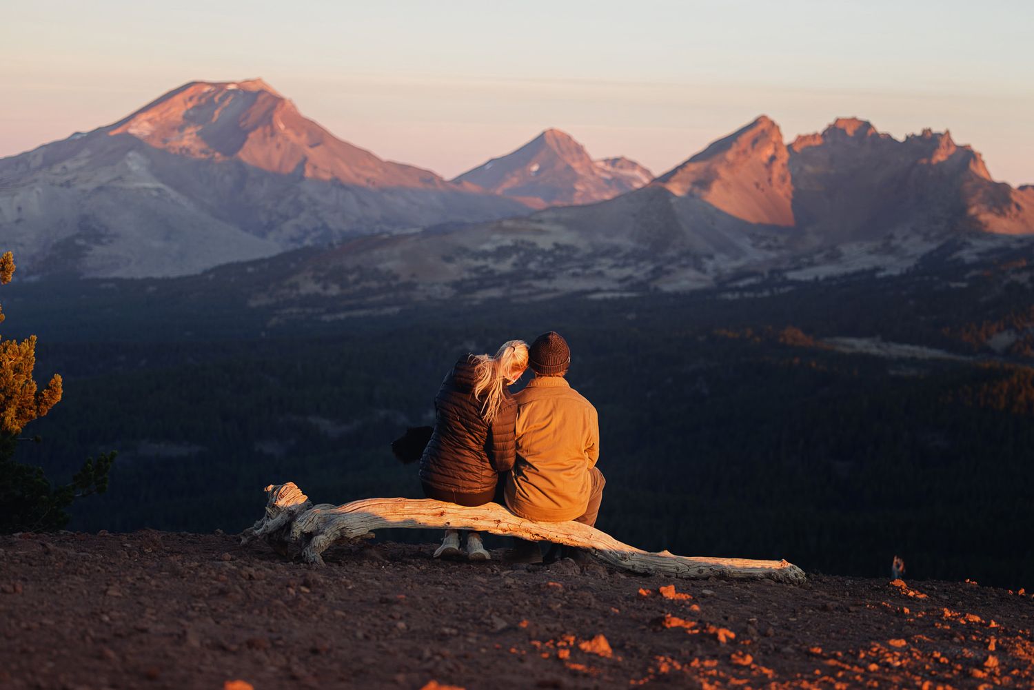 Two people sit together on a weathered log overlooking rugged mountain peaks glowing in warm golden sunset light.