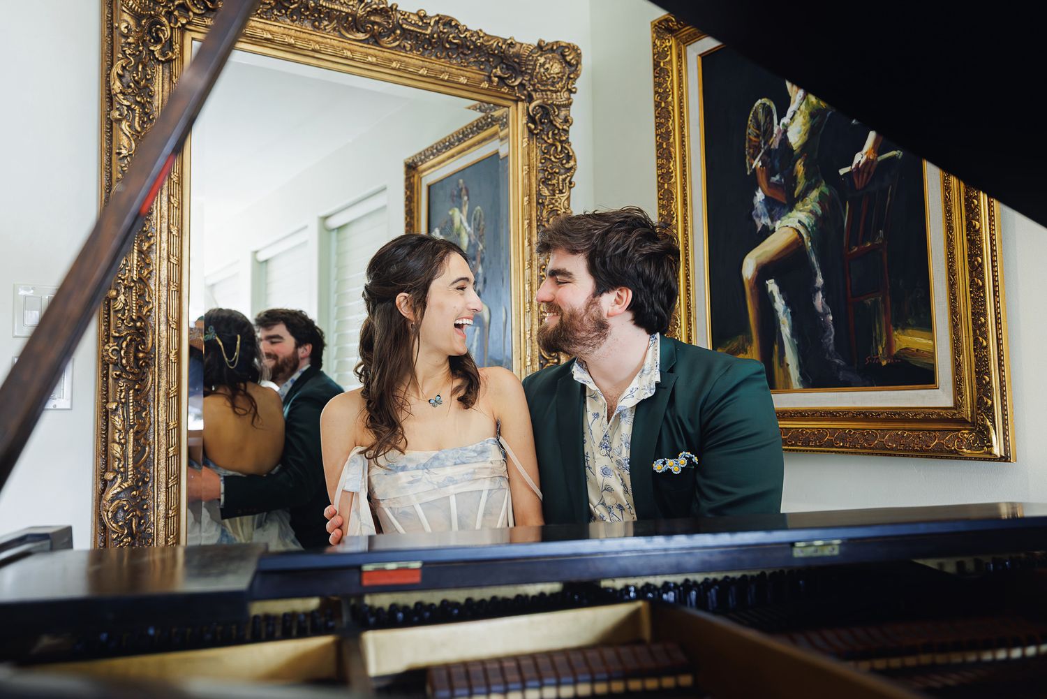 Couple smiling at each other while sitting at a grand piano, with gold-framed artwork on the wall behind them.