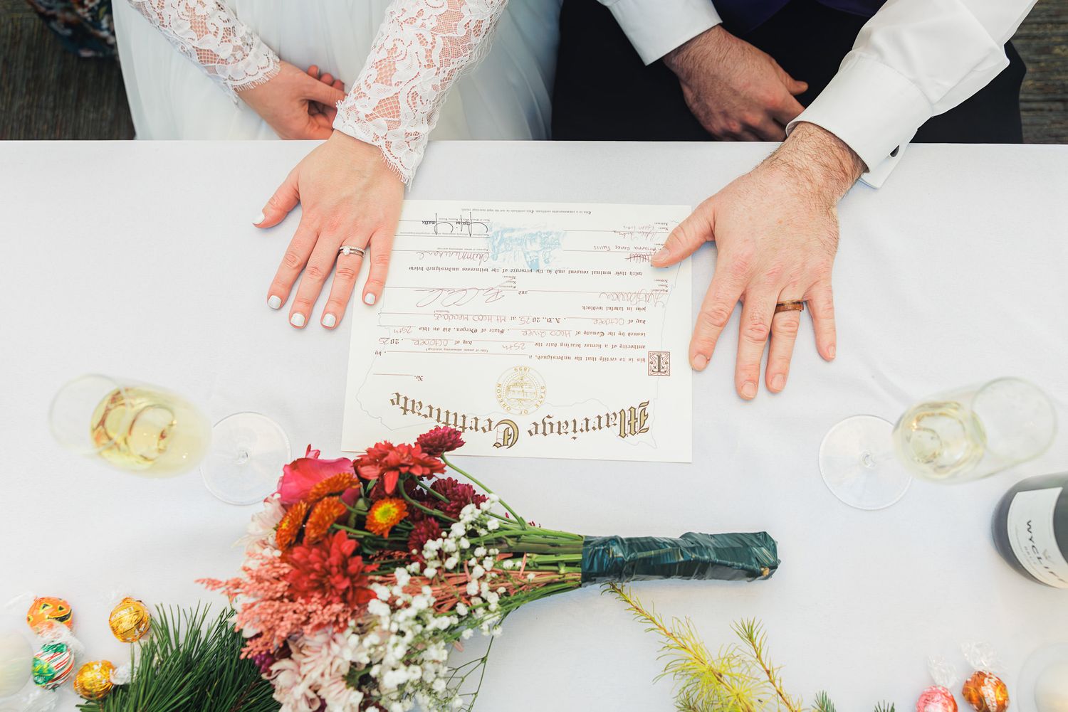 Wedding certificate signing with hands, rings and colorful floral bouquet on white table.