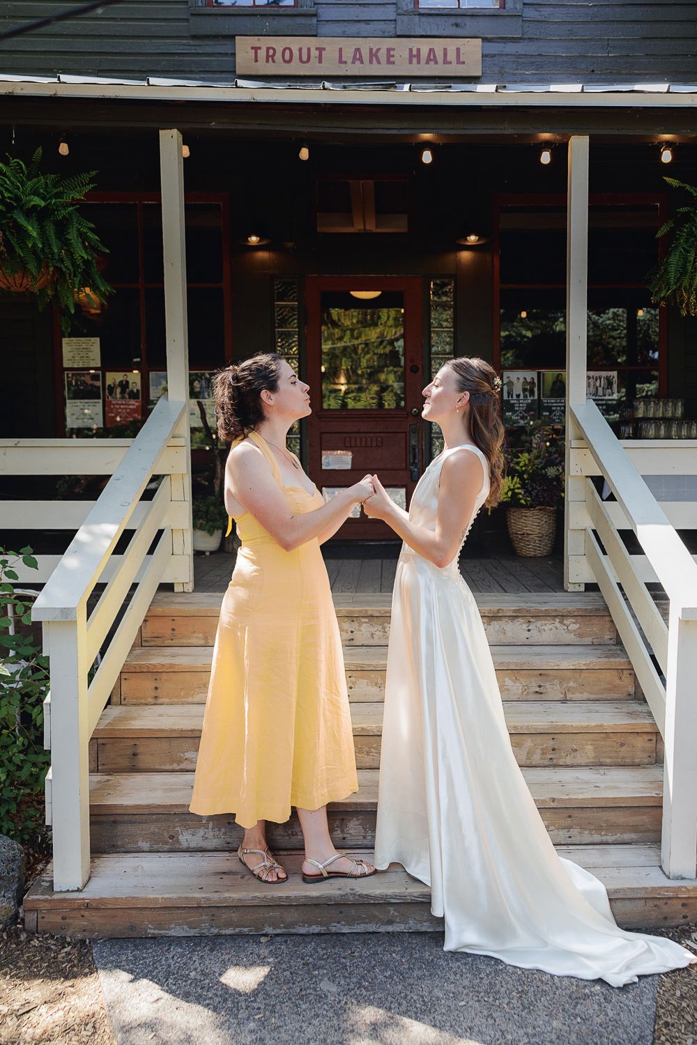 Two people in formal dresses share a joyful moment on wooden steps outside Trout Lake Hall.
