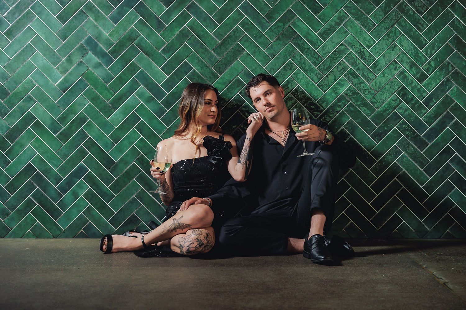 A couple sits together on the floor against a green herringbone tile wall, sharing a laugh in a casual pose.