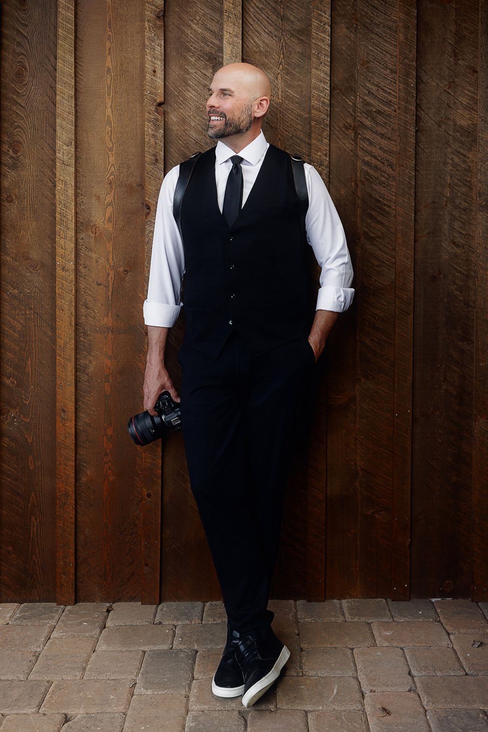 Professional photographer in formal black vest and tie holds camera while posing against rustic wooden wall.