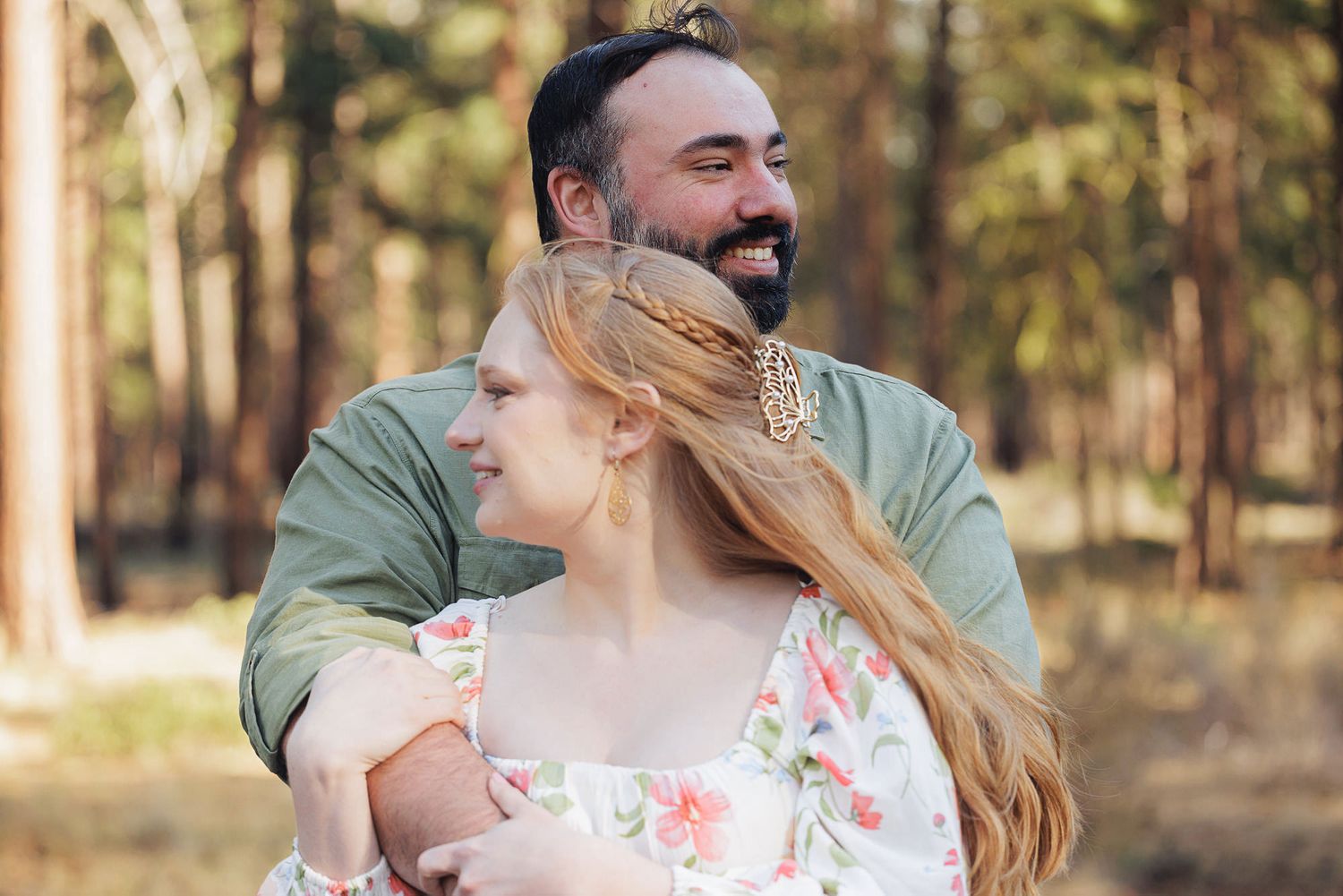 Couple embracing outdoors in a forest, woman in floral dress with long red hair, man in green shirt smiling.