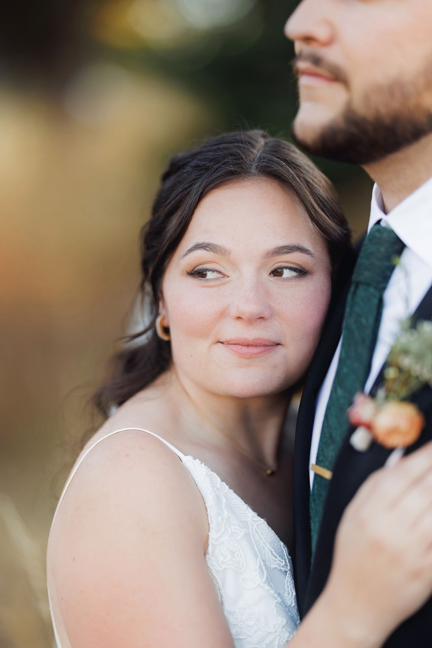 Bride in white lace dress smiles softly, leaning against groom in dark suit with green floral boutonniere.