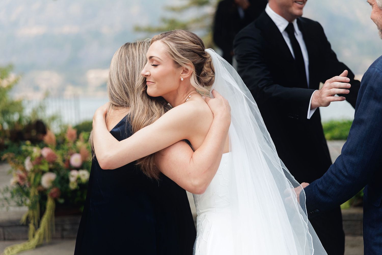 Bride in white gown and veil sharing a warm hug with a woman in black dress at an outdoor wedding ceremony.