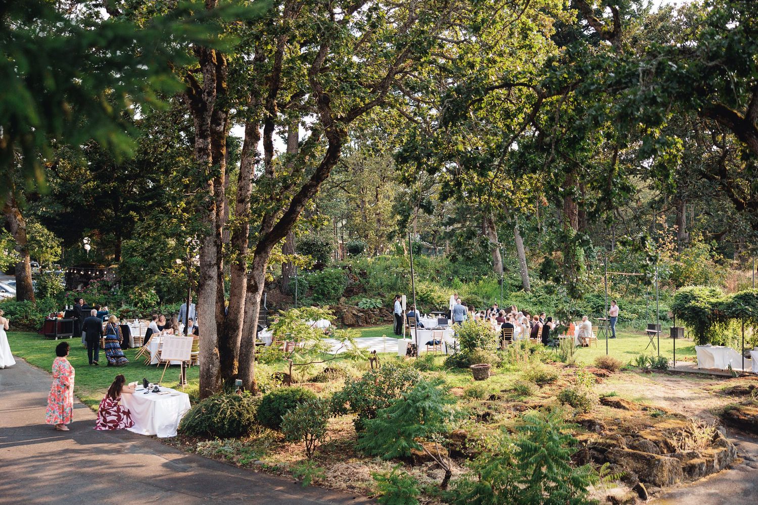 Outdoor garden wedding reception with guests mingling among lush trees and manicured hedges on a sunny day.