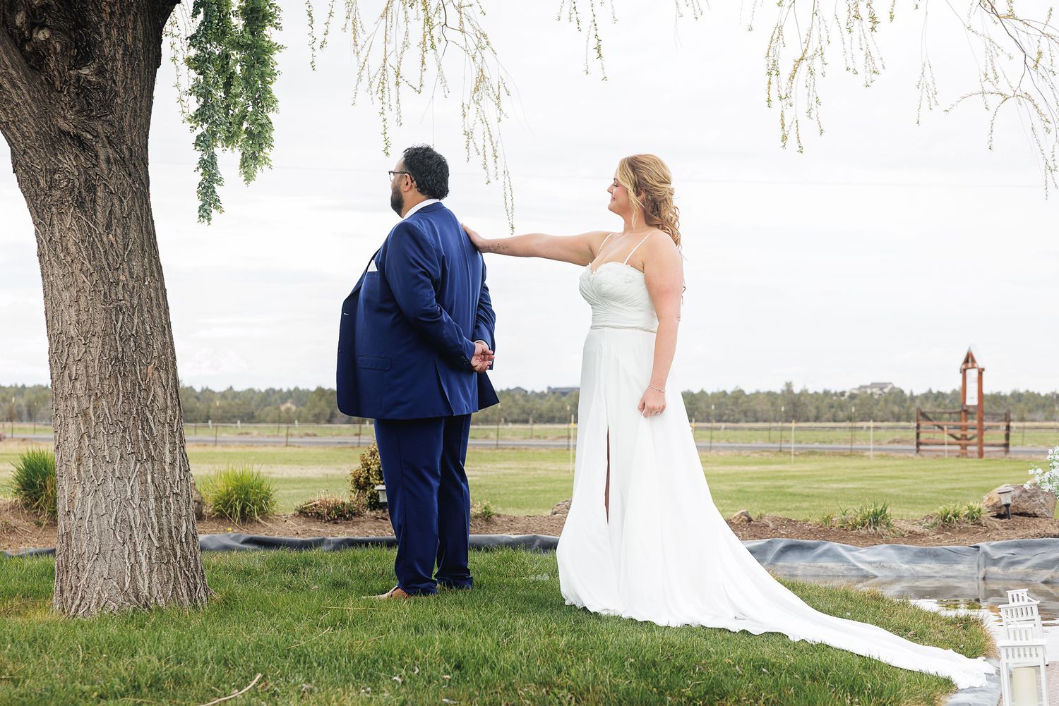 Bride in white gown taps groom in navy suit on shoulder for first look moment under willow tree at outdoor wedding venue.