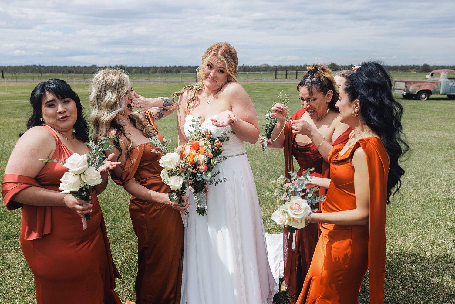 Bride in white gown with four bridesmaids in rust orange dresses holding bouquets outdoors at a wedding venue.