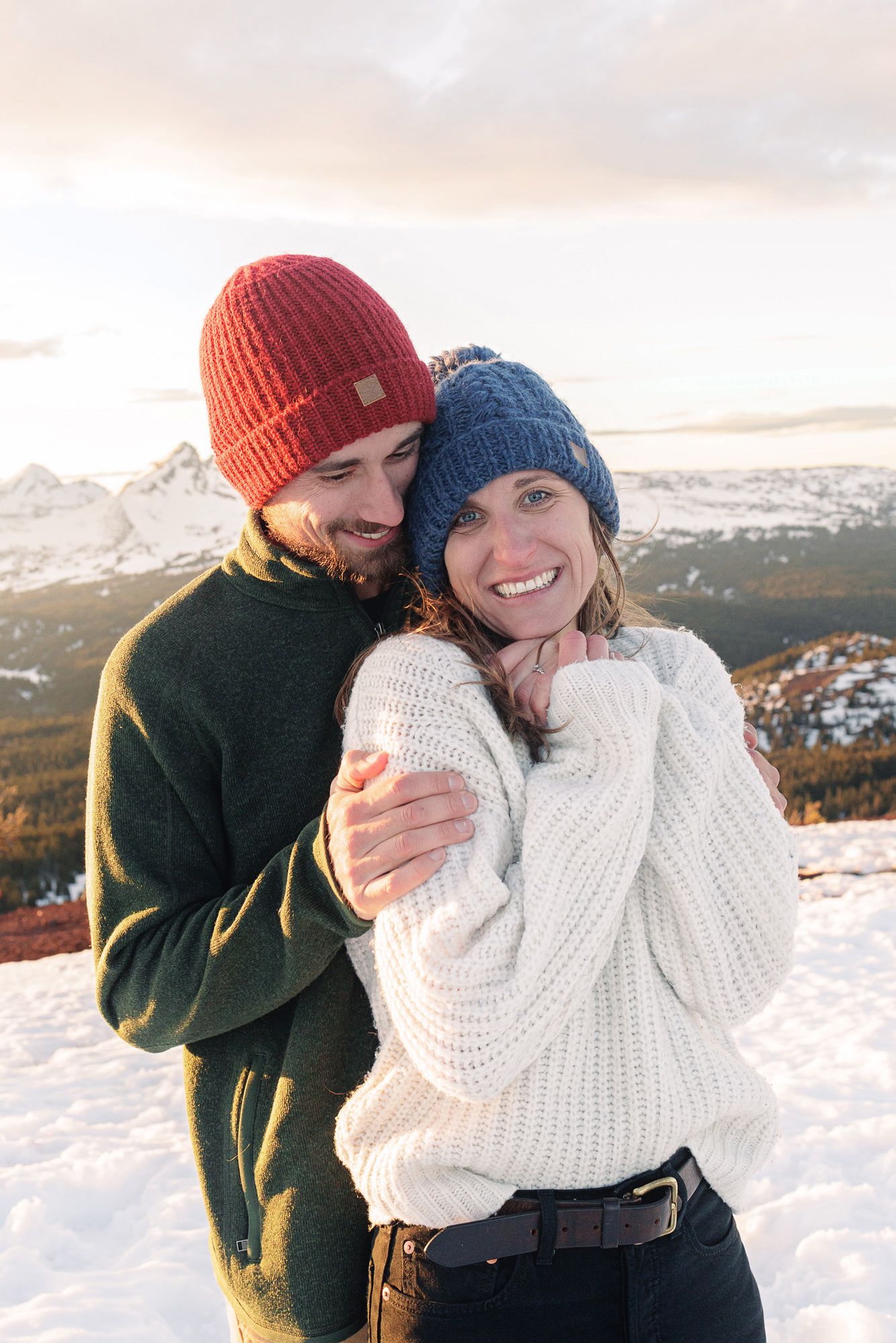 Couple in winter hats embracing on snowy mountain, woman in white sweater smiling, scenic alpine backdrop at sunset.