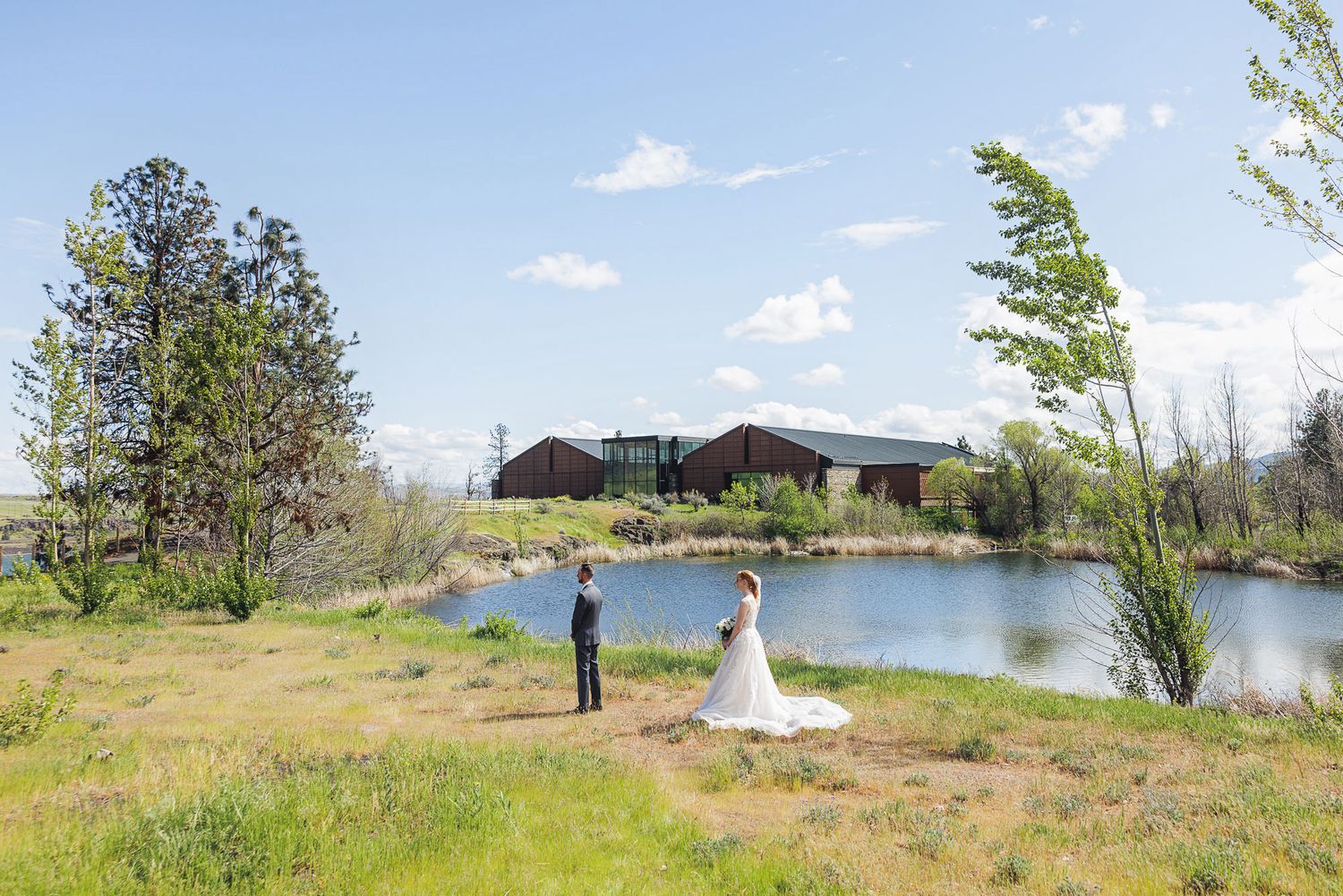 Bride and groom share a romantic first look by a serene pond with a rustic barn venue in the background on a sunny day.
