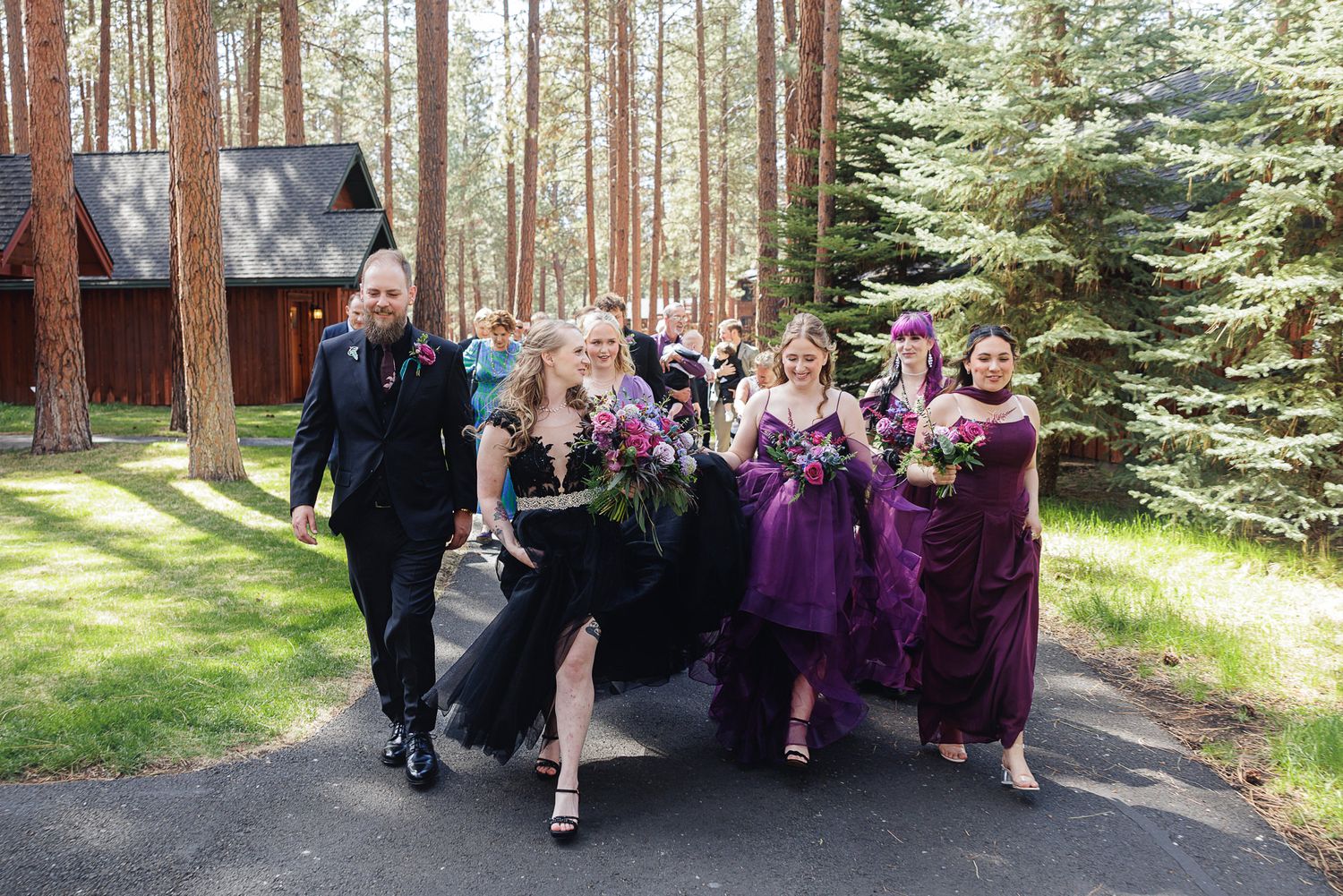 Wedding party walks outdoors among tall pine trees, bride in black gown with colorful bridesmaids in purple dresses.