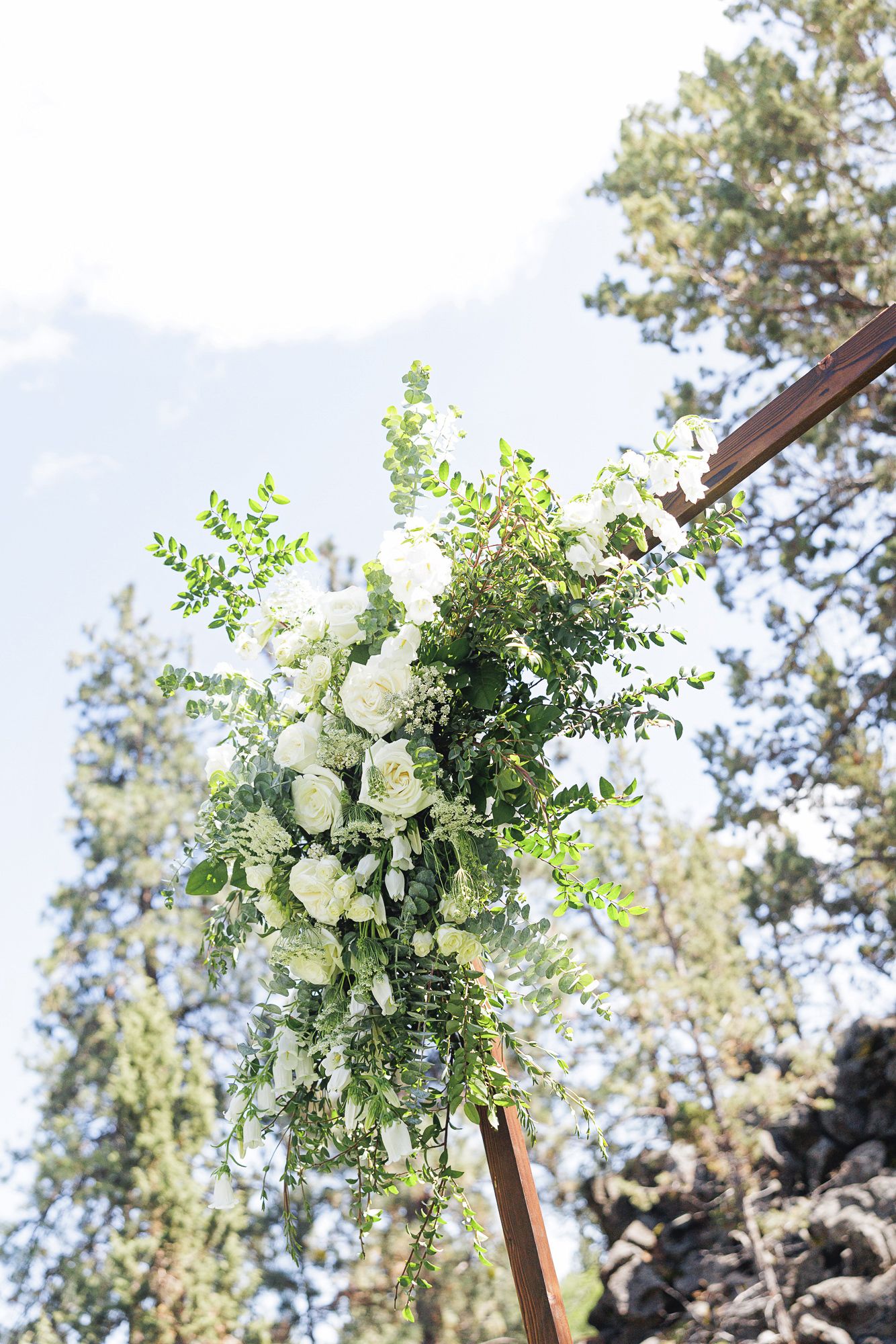 White floral arrangement with lush greenery on a wooden wedding arch, set against tall pine trees and a bright sky.