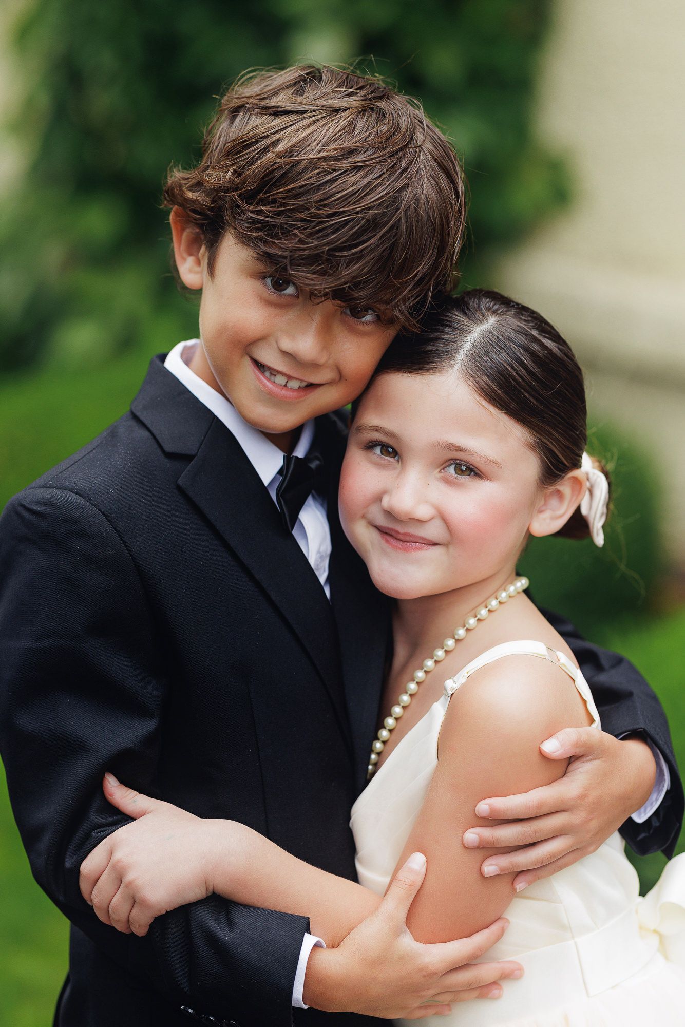 Two children in formal attire embrace outdoors, boy in black suit hugs girl in white dress with pearl necklace.
