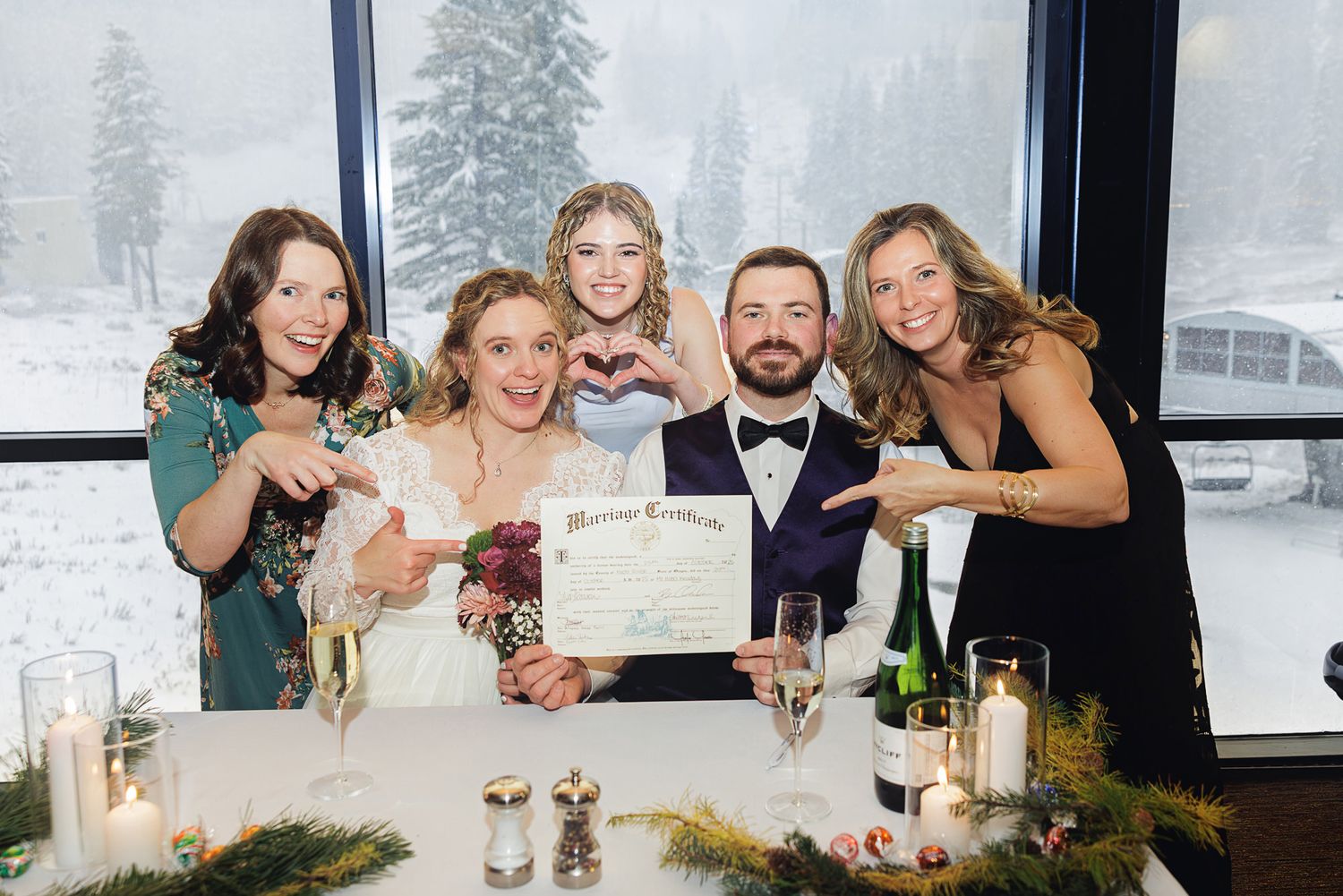 Wedding party celebrates at reception table with wine glasses and snowy winter scene visible through windows.