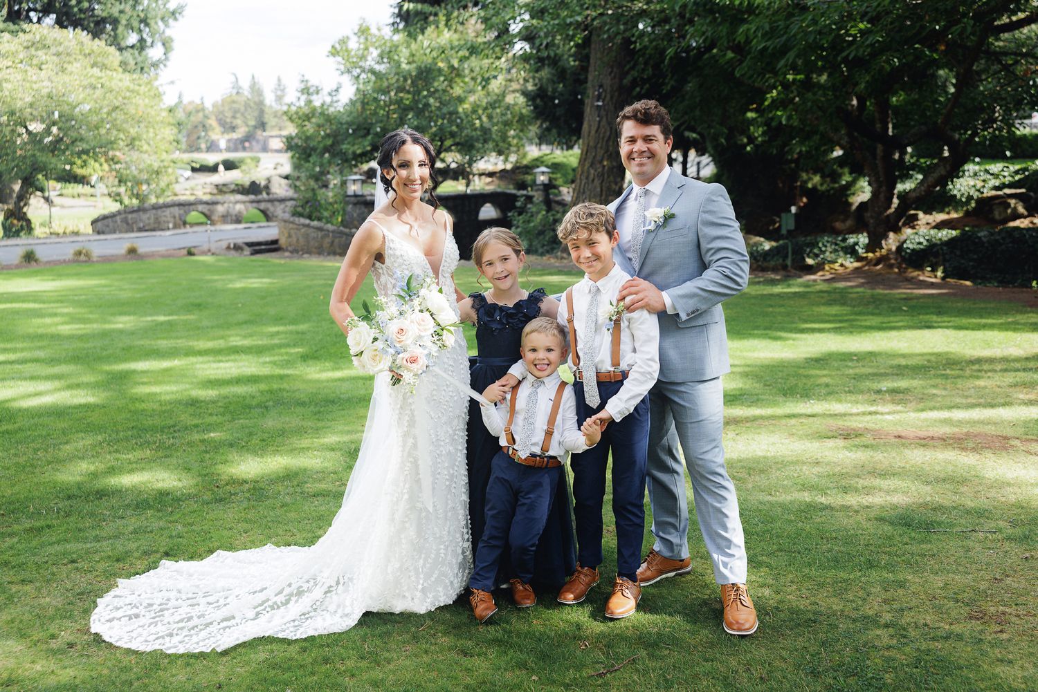 A wedding party poses together on a lush green lawn during a sunny outdoor celebration.