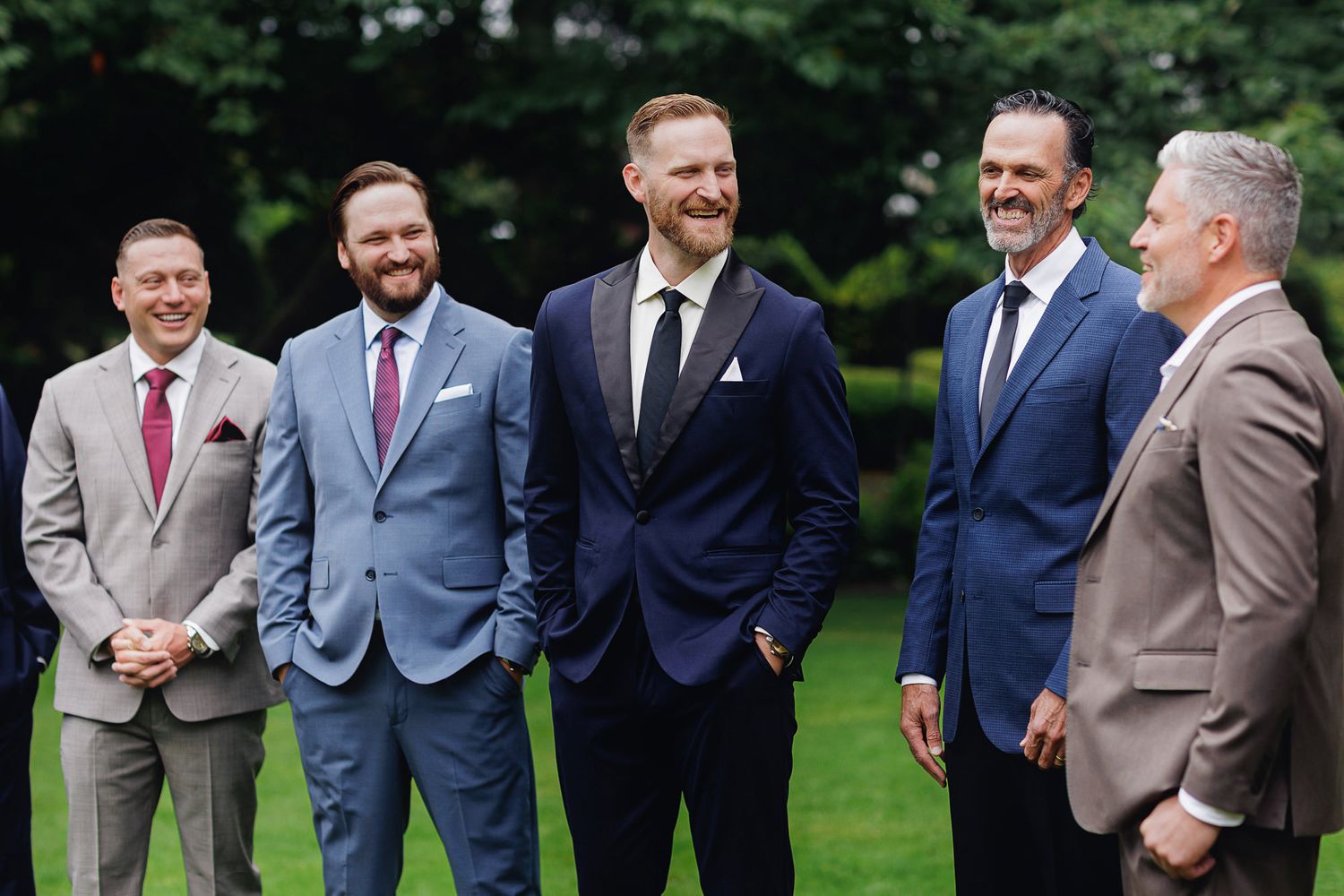 Five men in suits smiling and laughing together outdoors on a green lawn at a formal event.