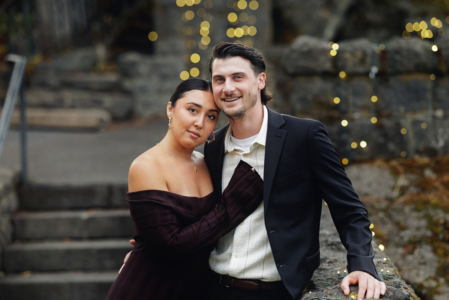 Couple posing together in formal attire with bokeh lights in background, woman in burgundy off-shoulder dress.
