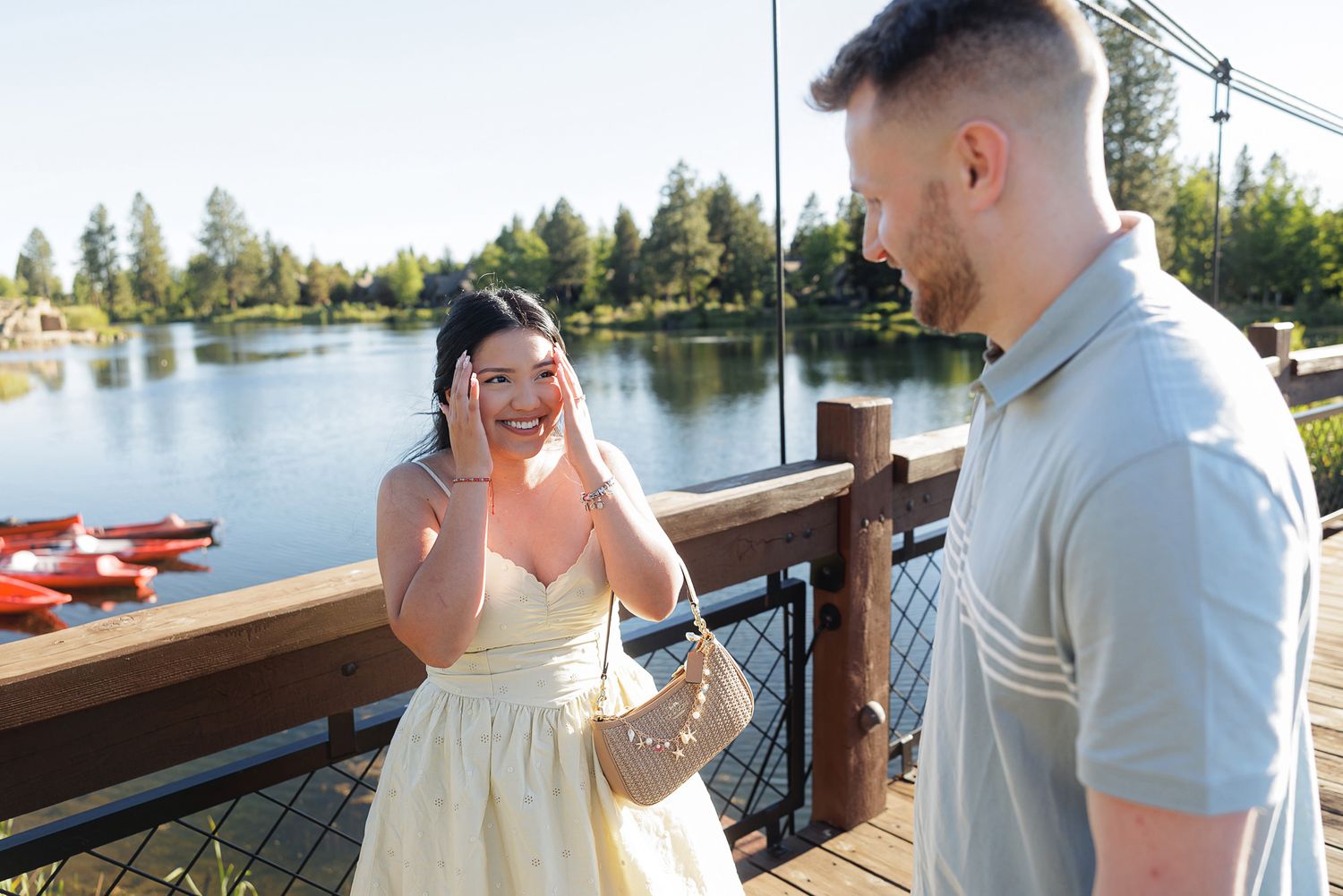 A joyful woman reacts emotionally on a lakeside dock as a man presents her with an engagement ring outdoors.