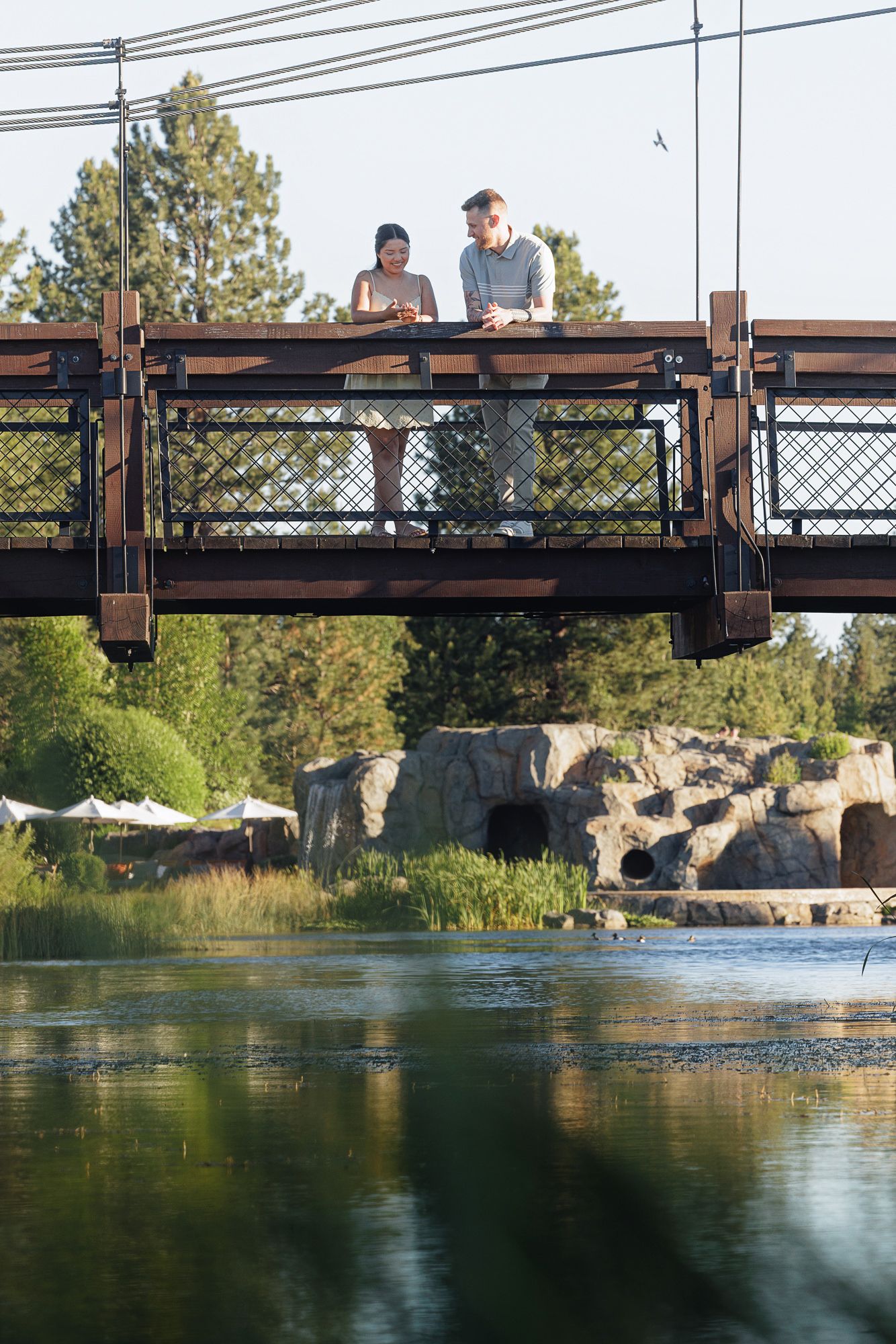 Couple standing on a wooden suspension bridge over a calm river surrounded by lush green trees and rocky landscape.