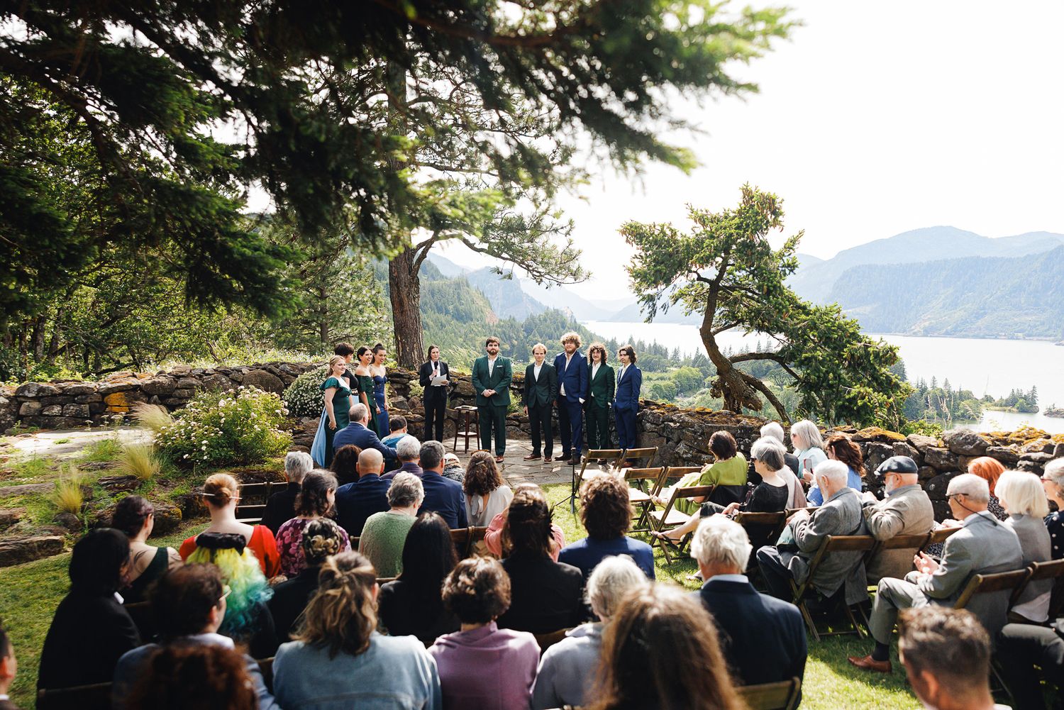 Outdoor wedding ceremony with guests seated on chairs, wedding party in teal, overlooking a scenic mountain river valley.