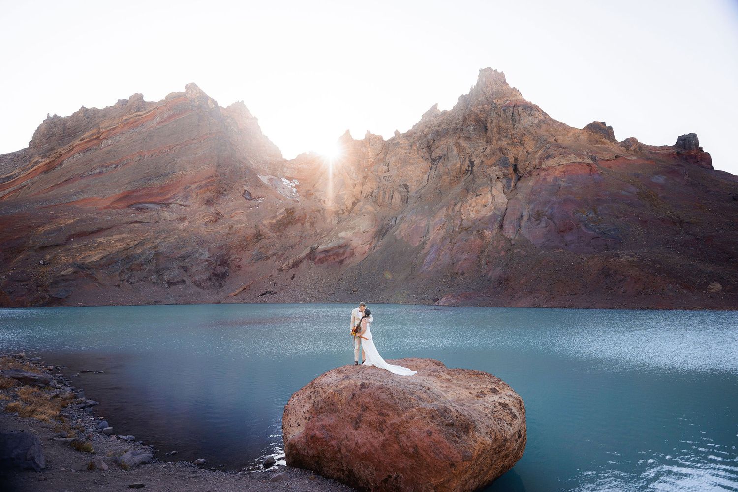 Couple embraces on a large rock in a turquoise alpine lake, with rugged red mountain peaks and glowing sunlight behind them.
