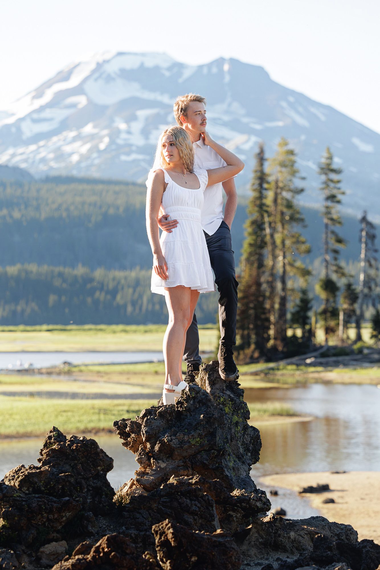 Couple standing on volcanic rocks with snowy mountain backdrop, woman in white dress embracing man in casual attire.