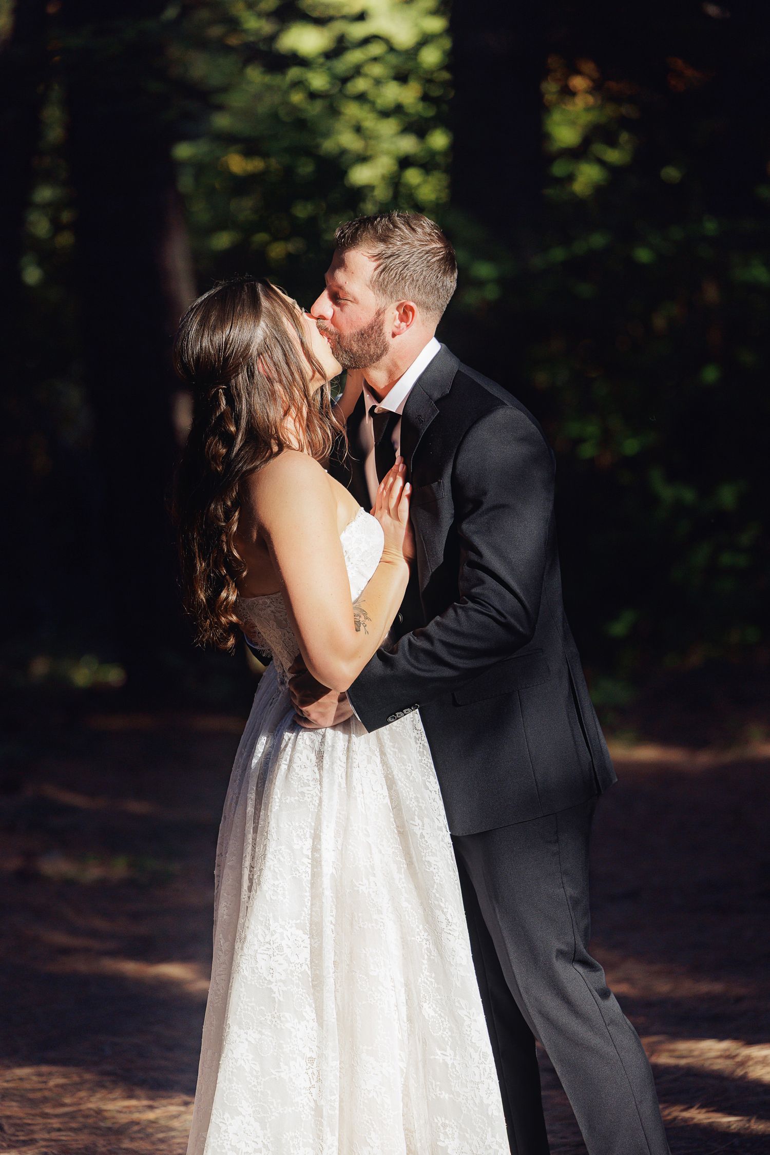 Bride and groom share a romantic kiss outdoors in a sunlit forest, she in a white gown, he in a dark suit.