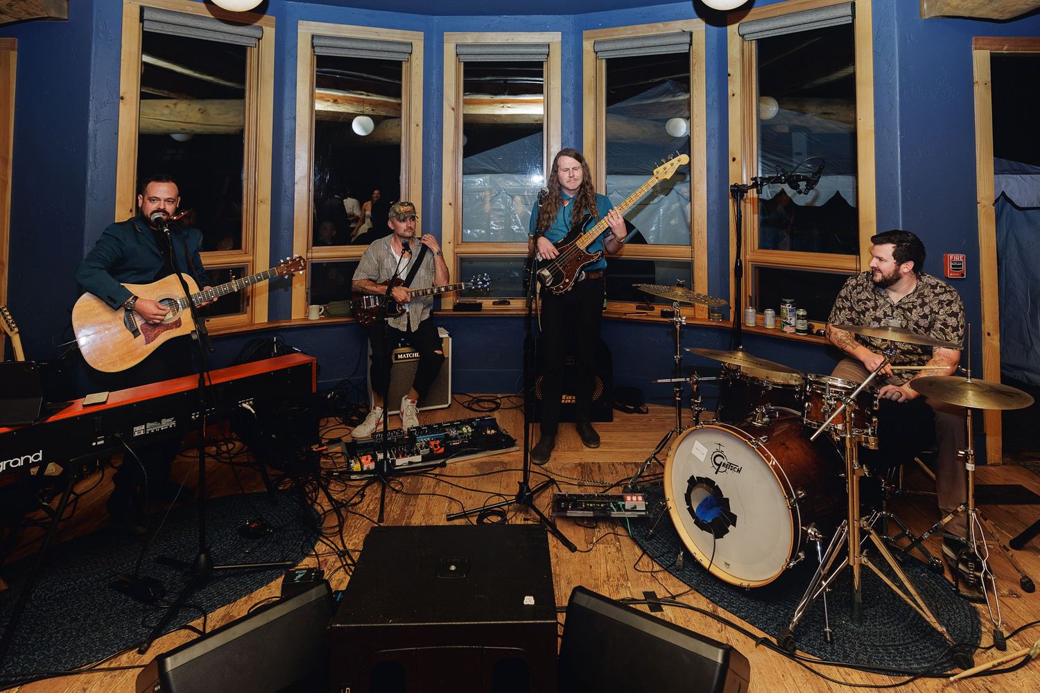 Four-piece band performing in a blue recording studio with guitars, keyboard, and drums on hardwood floors.