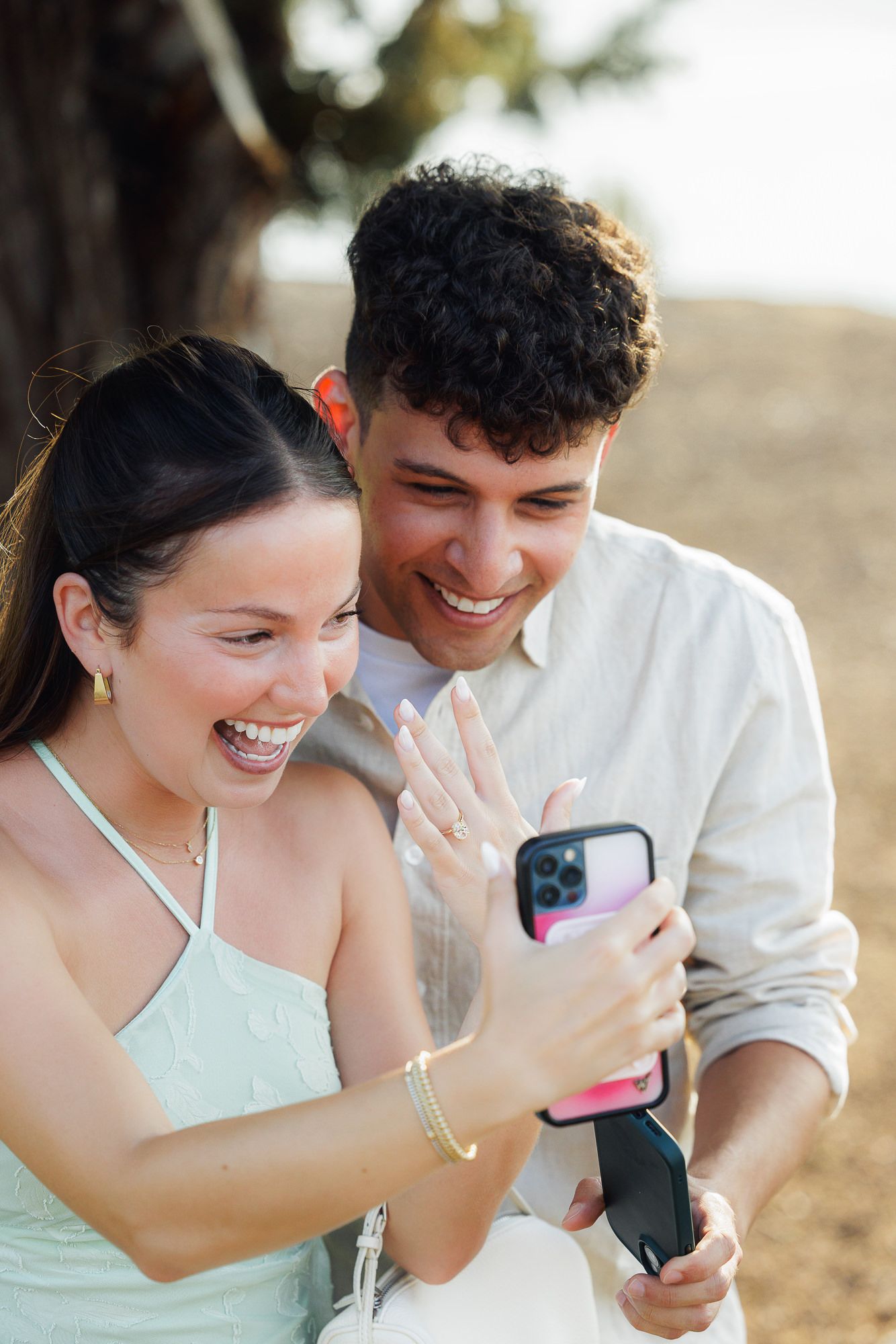 Happy couple smiling at engagement ring on her finger while video calling on a pink iPhone outdoors.