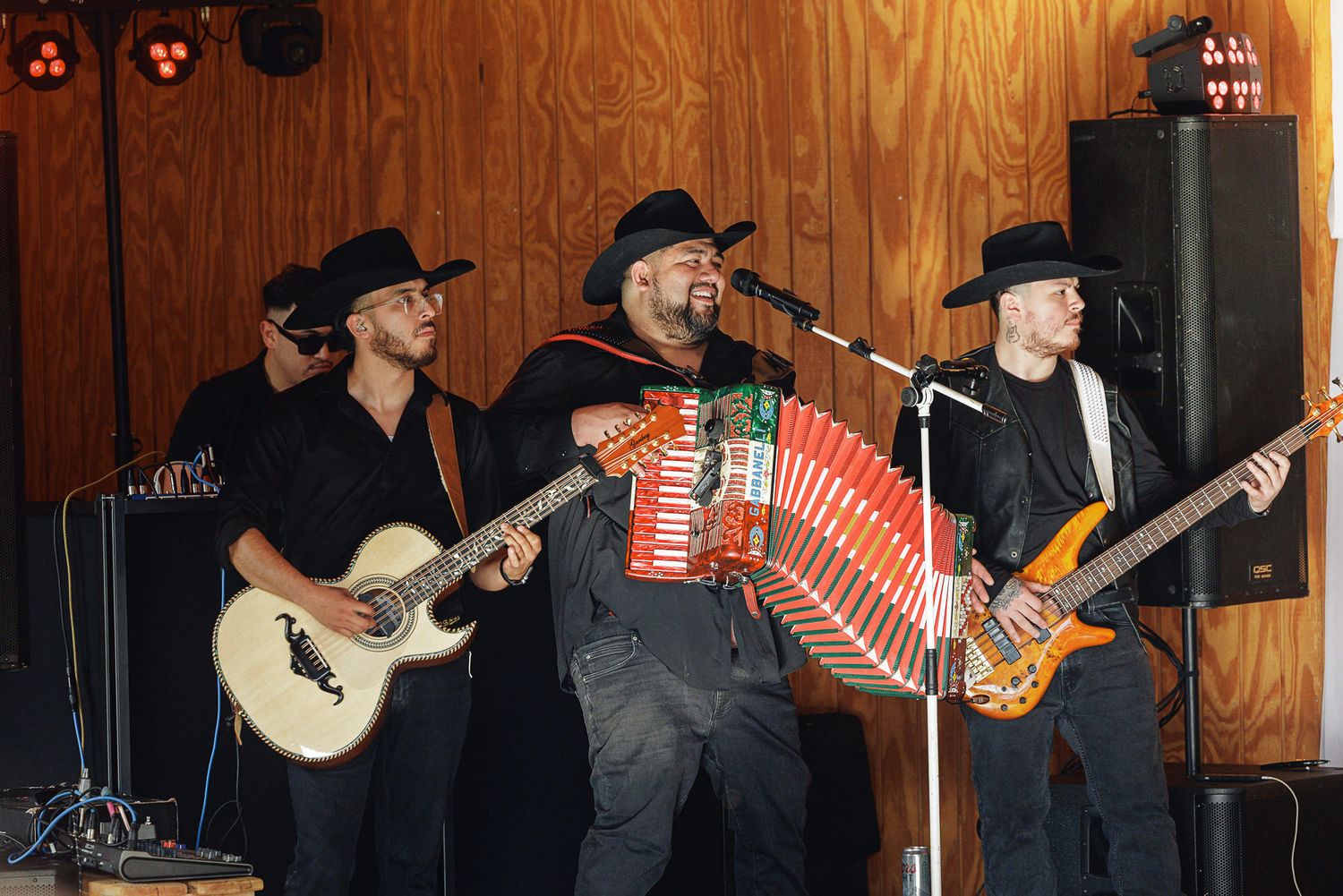 Three musicians perform on stage, featuring accordion, acoustic and bass guitars, all wearing black cowboy hats.