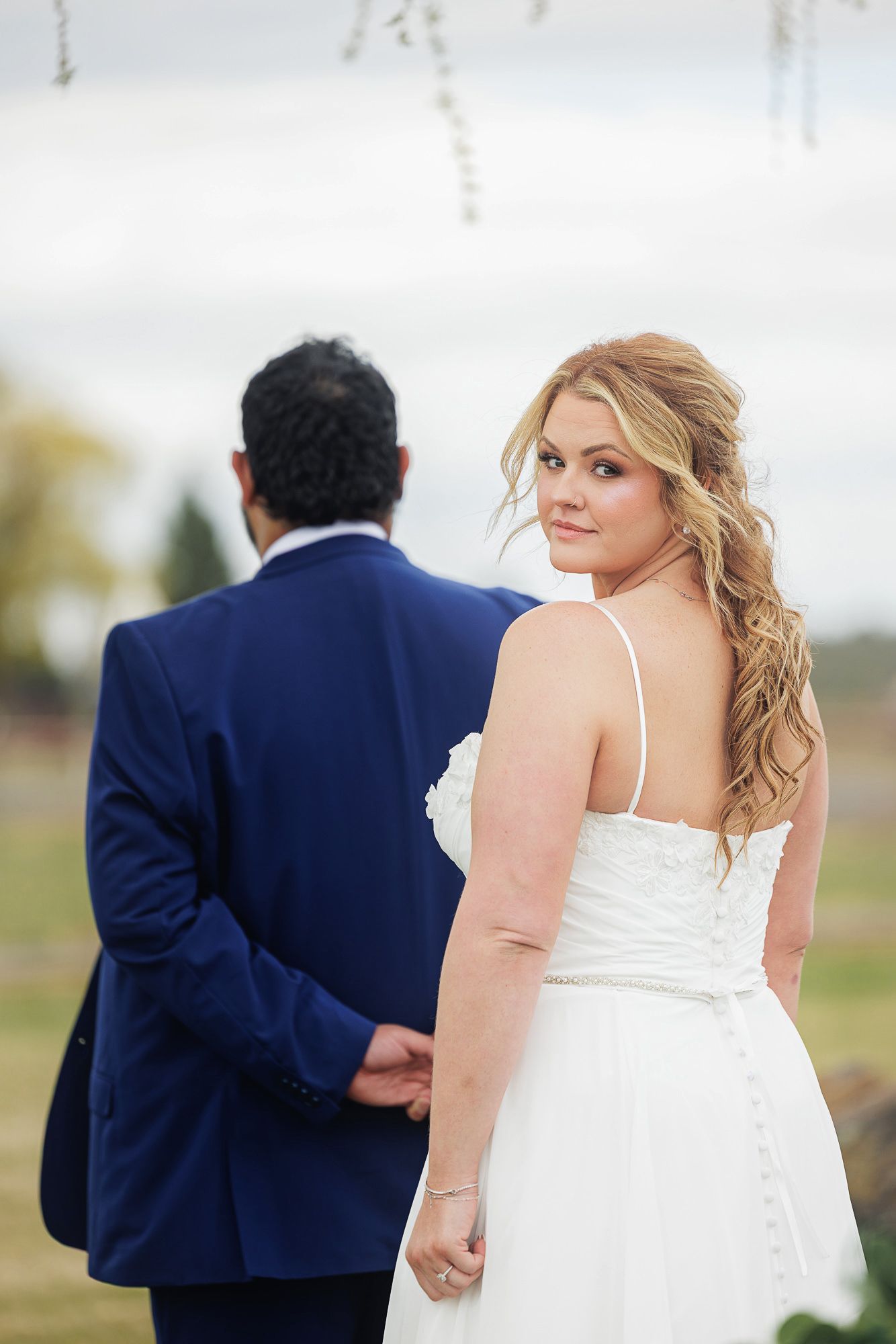Bride in white gown looks back at camera while holding hands with groom in navy suit outdoors.