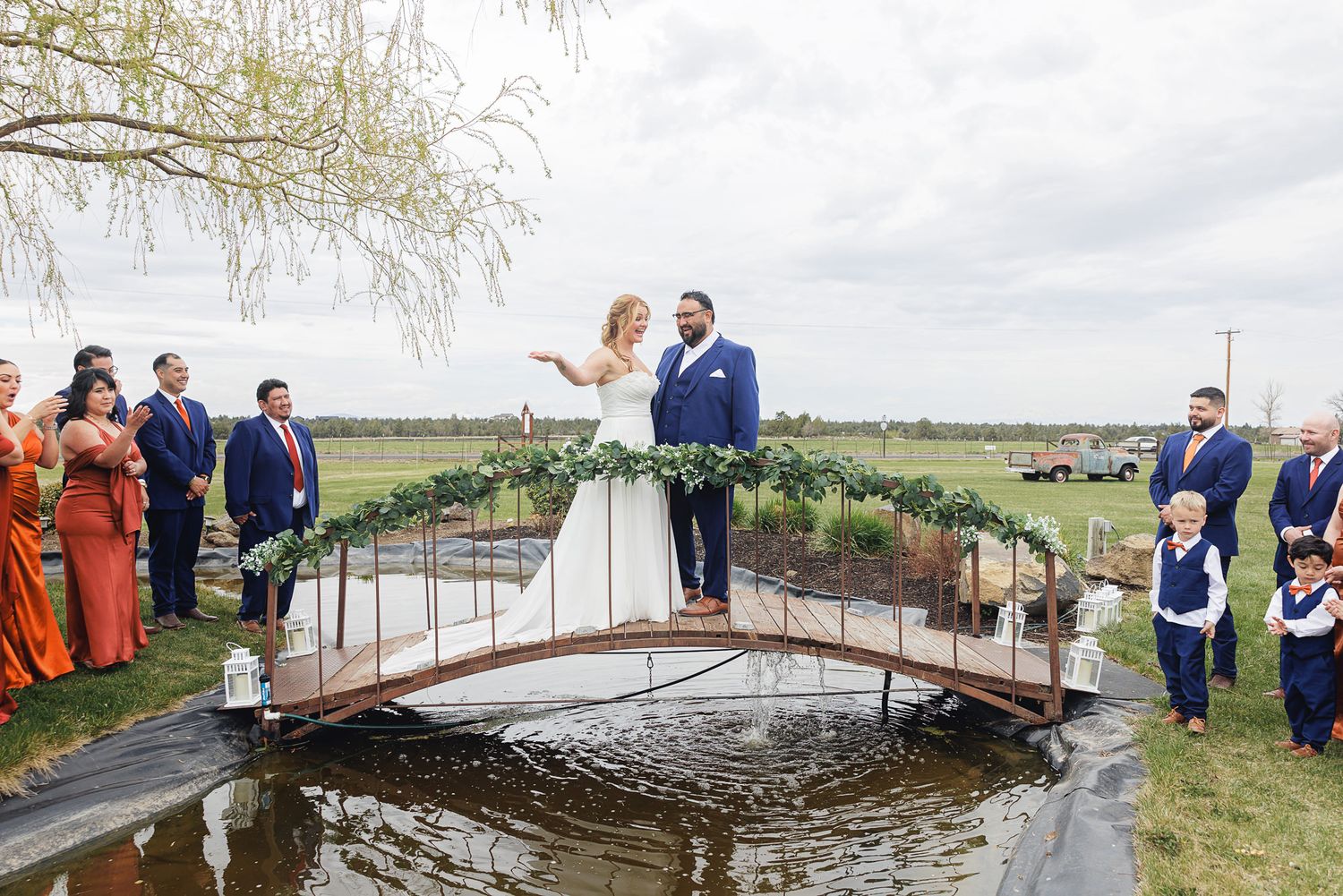 Bride and groom exchange vows on a wooden bridge over water at an outdoor wedding ceremony with guests watching.