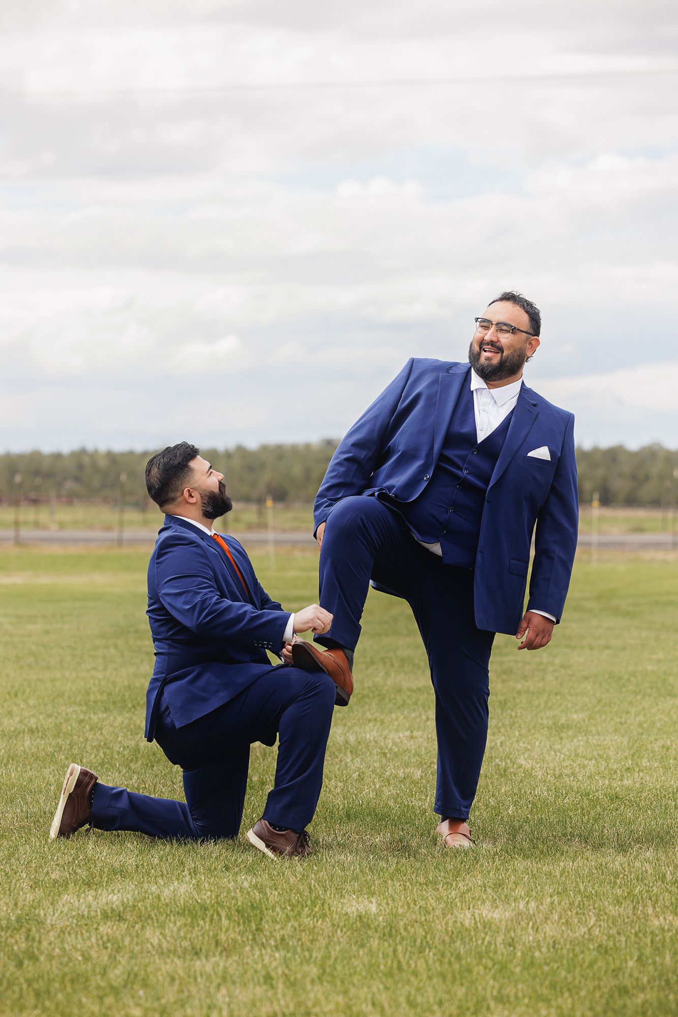 Two grooms in matching blue suits share a playful moment on a green lawn during their wedding day.