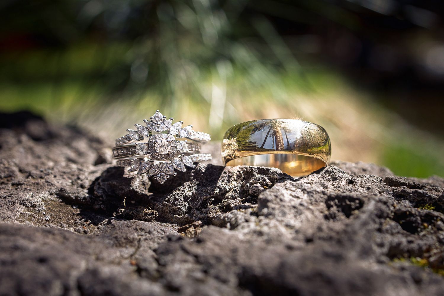 Close-up of diamond engagement ring and gold wedding bands resting on dark rocky surface outdoors.