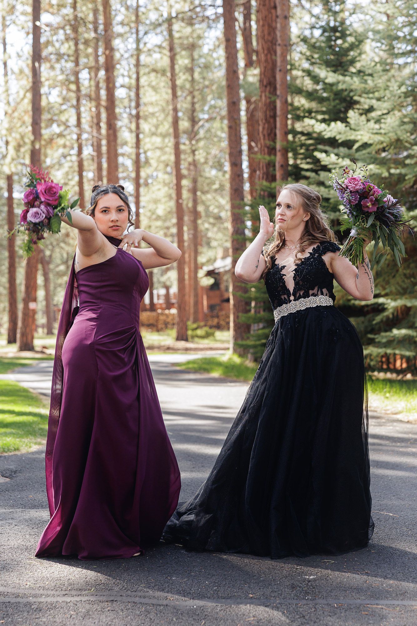 Two women in elegant gowns, one purple and one black, pose outdoors holding floral bouquets among tall pine trees.