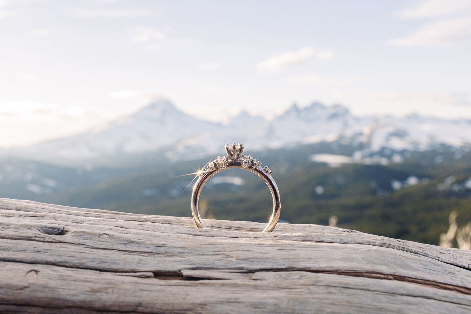 Gold diamond engagement ring displayed on a log with scenic mountain peaks and blue sky in the background.