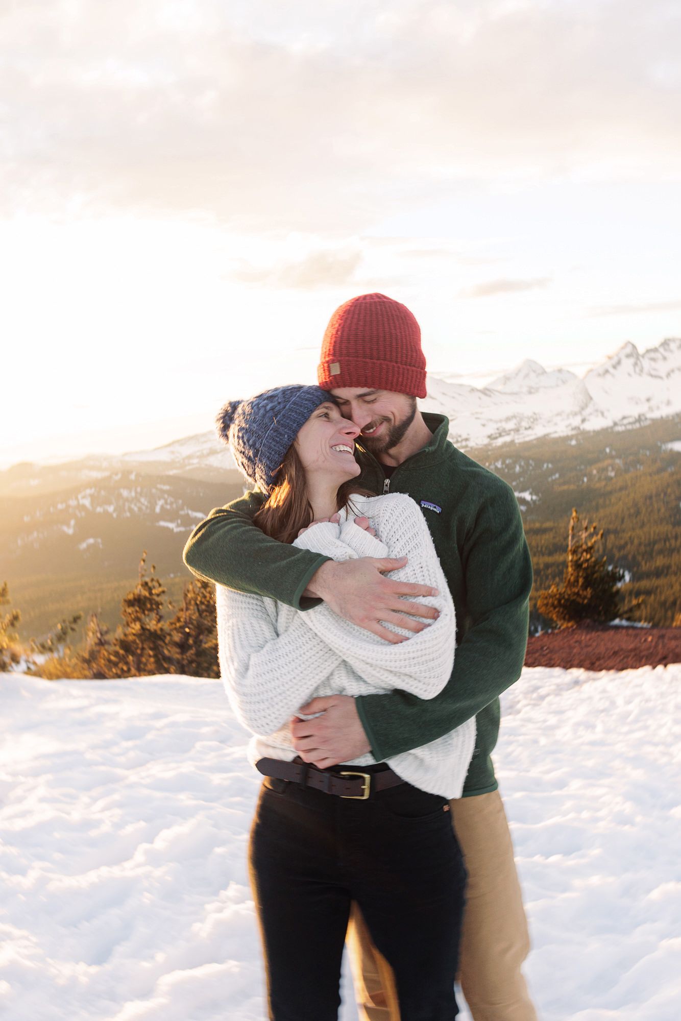 Couple embracing and smiling in snowy mountain landscape at golden hour, wearing winter hats and cozy sweaters.