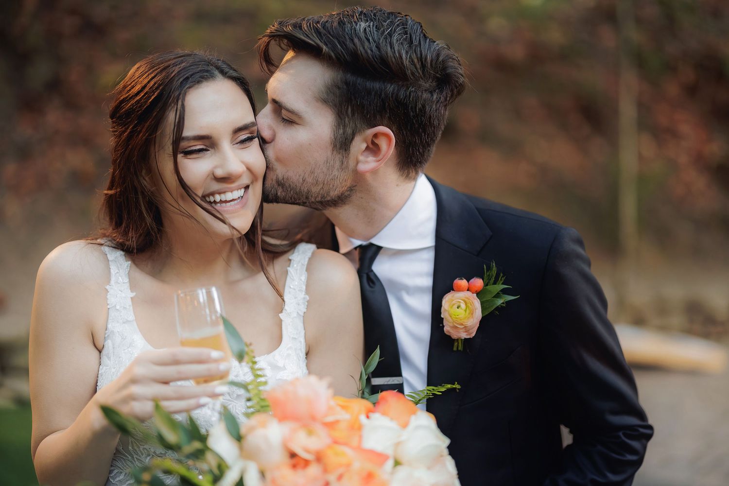 A tender newlywed moment as a bride holding a champagne flute receives a gentle kiss while holding a peach floral bouquet.