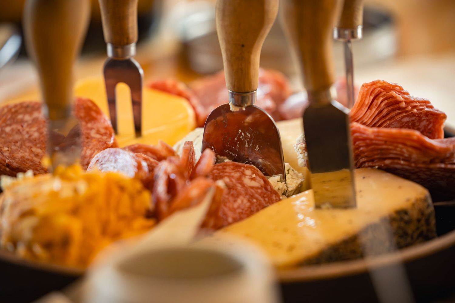 Serving utensils and forks dig into a charcuterie board filled with meats, cheeses and various appetizers.