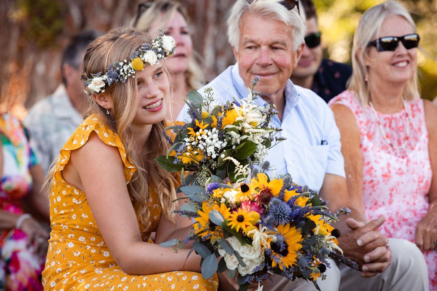 A person wearing a yellow polka dot dress holds a vibrant bouquet of sunflowers and wildflowers at an outdoor gathering.