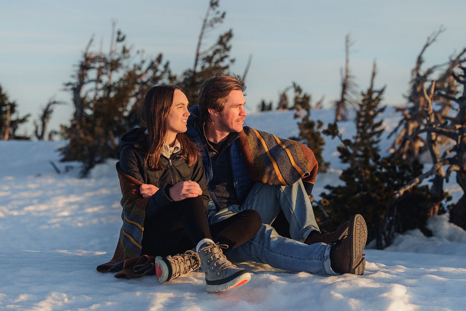 Two people sitting in snow at sunset, enjoying winter views of snowy evergreen trees and mountains.