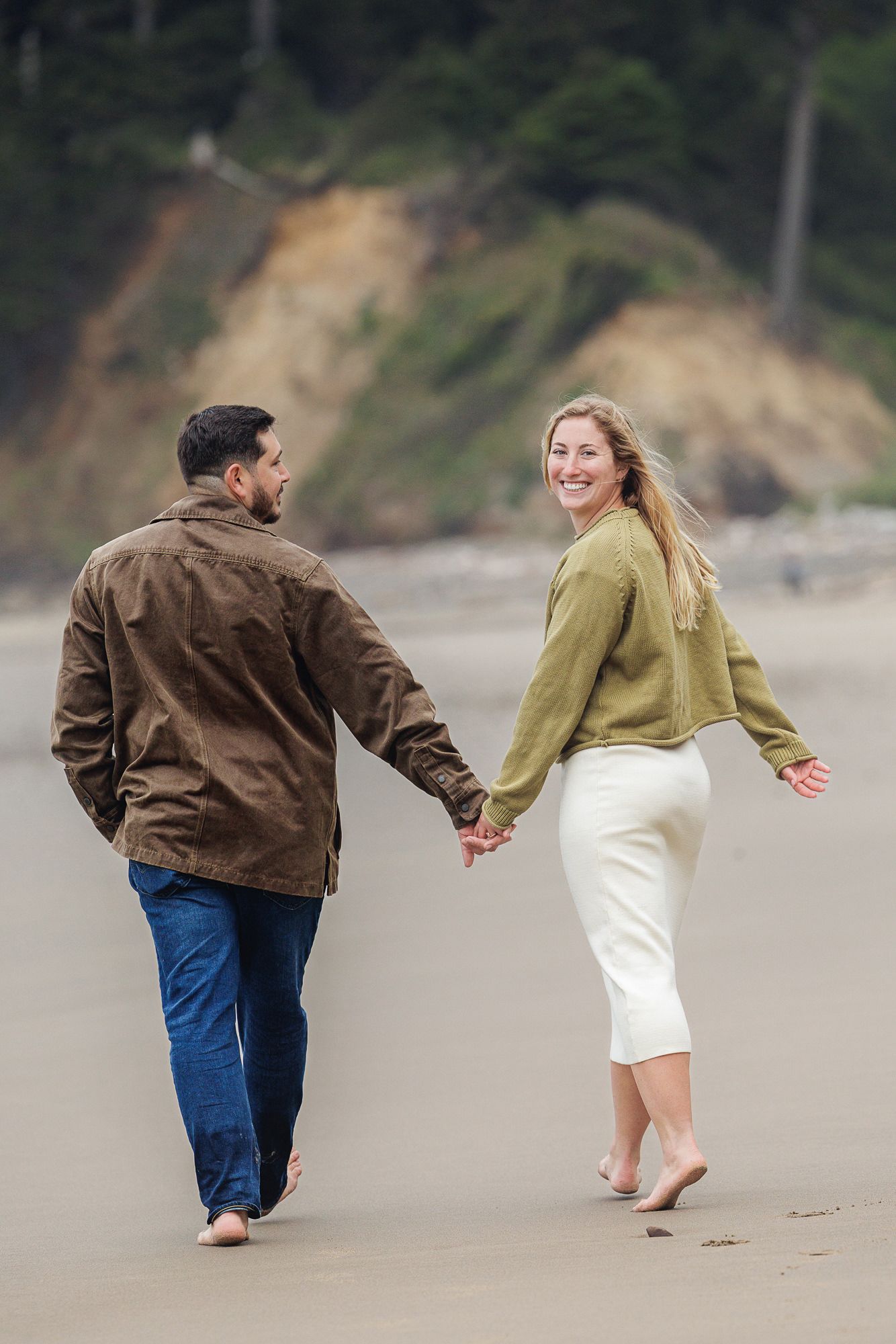 A smiling couple walks barefoot on a beach, holding hands, dressed in casual fall attire with coastal cliffs in the background.