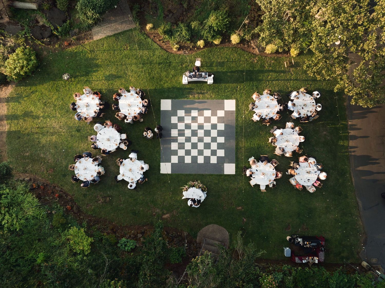 Aerial view of an outdoor wedding reception with round tables surrounding a giant chess board on green lawn.