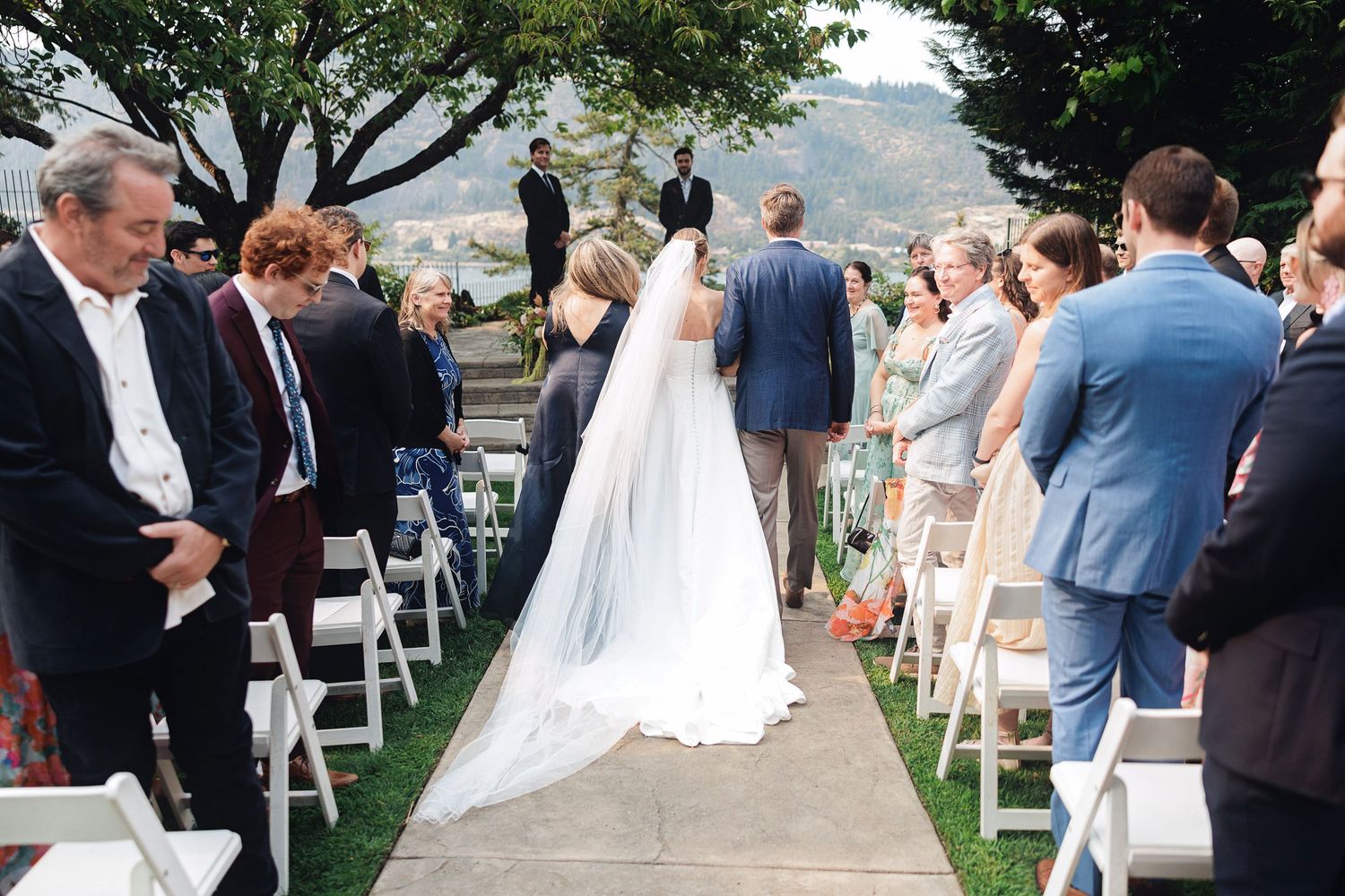 Bride walks down outdoor aisle in white gown and veil, guests seated in white chairs, officiant stands at altar.
