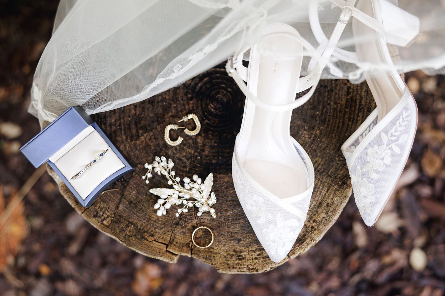 Wedding accessories flat lay featuring lace heels, diamond rings, gold band, pearl earrings, and floral hair piece on wood stump.