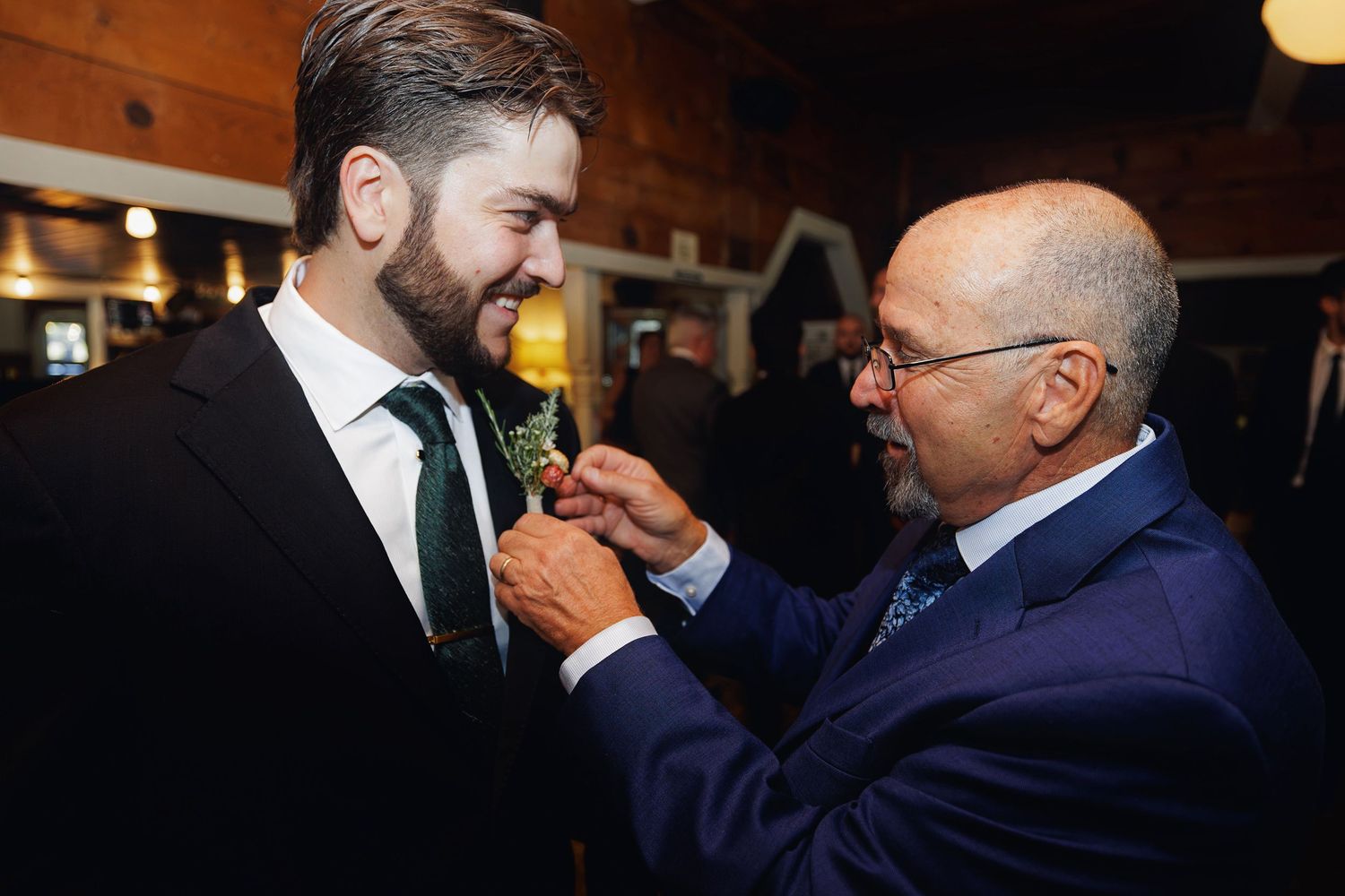 An older man pins a boutonniere on a younger bearded man in a suit at a formal wedding event.