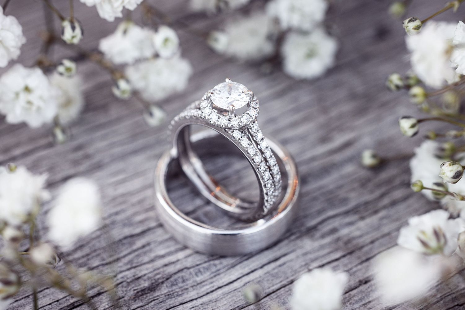 Diamond engagement ring and wedding band surrounded by delicate white flowers on rustic wooden background.